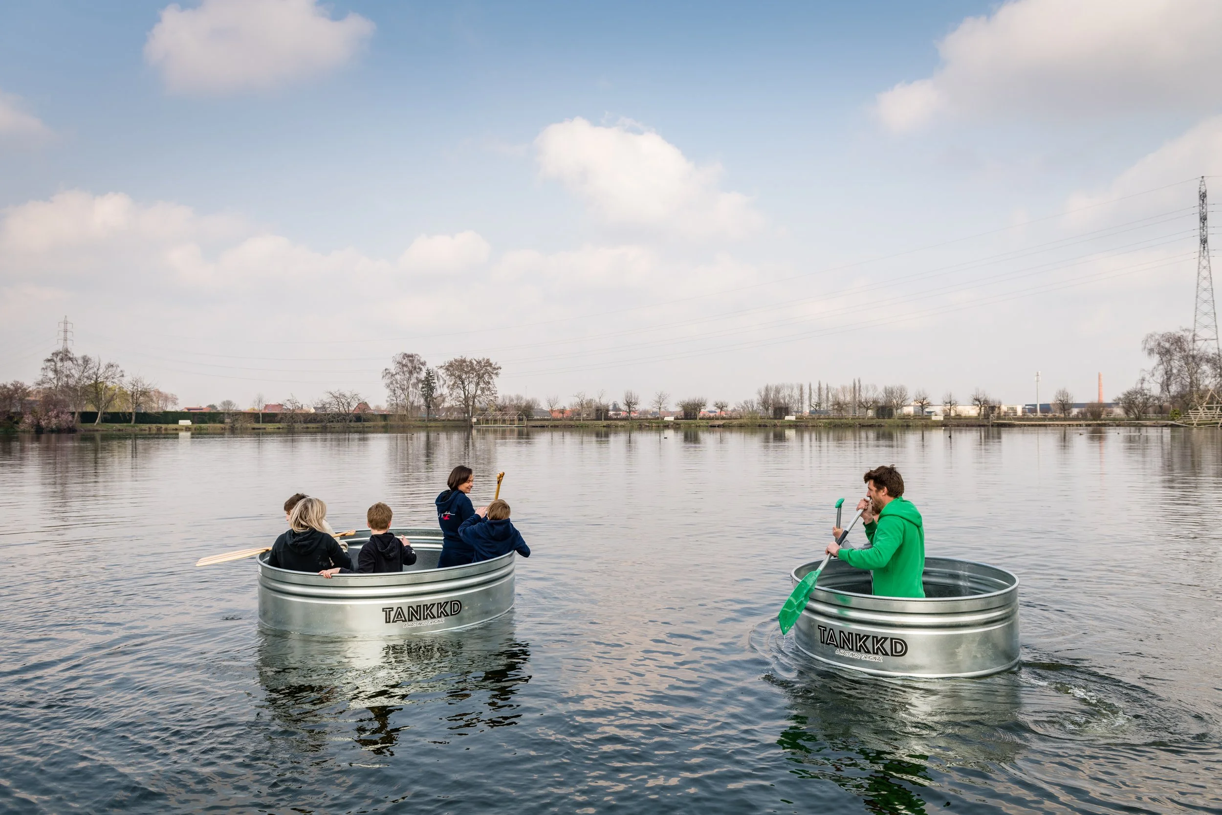 Two round Stock Tanks labeled TANKKD floating on a calm lake, each with multiple people paddling and enjoying the day, with a partly cloudy sky and distant trees and power lines in the background.