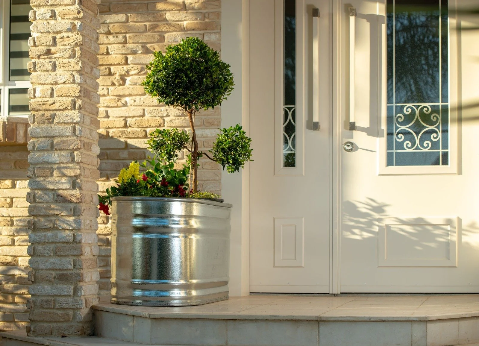 Close-up of a front porch with a metal flower pot containing a small tree and colorful flowers, a brick wall on the left, a white door with glass panel and decorative grille on the right, and a shadow cast by a plant.
