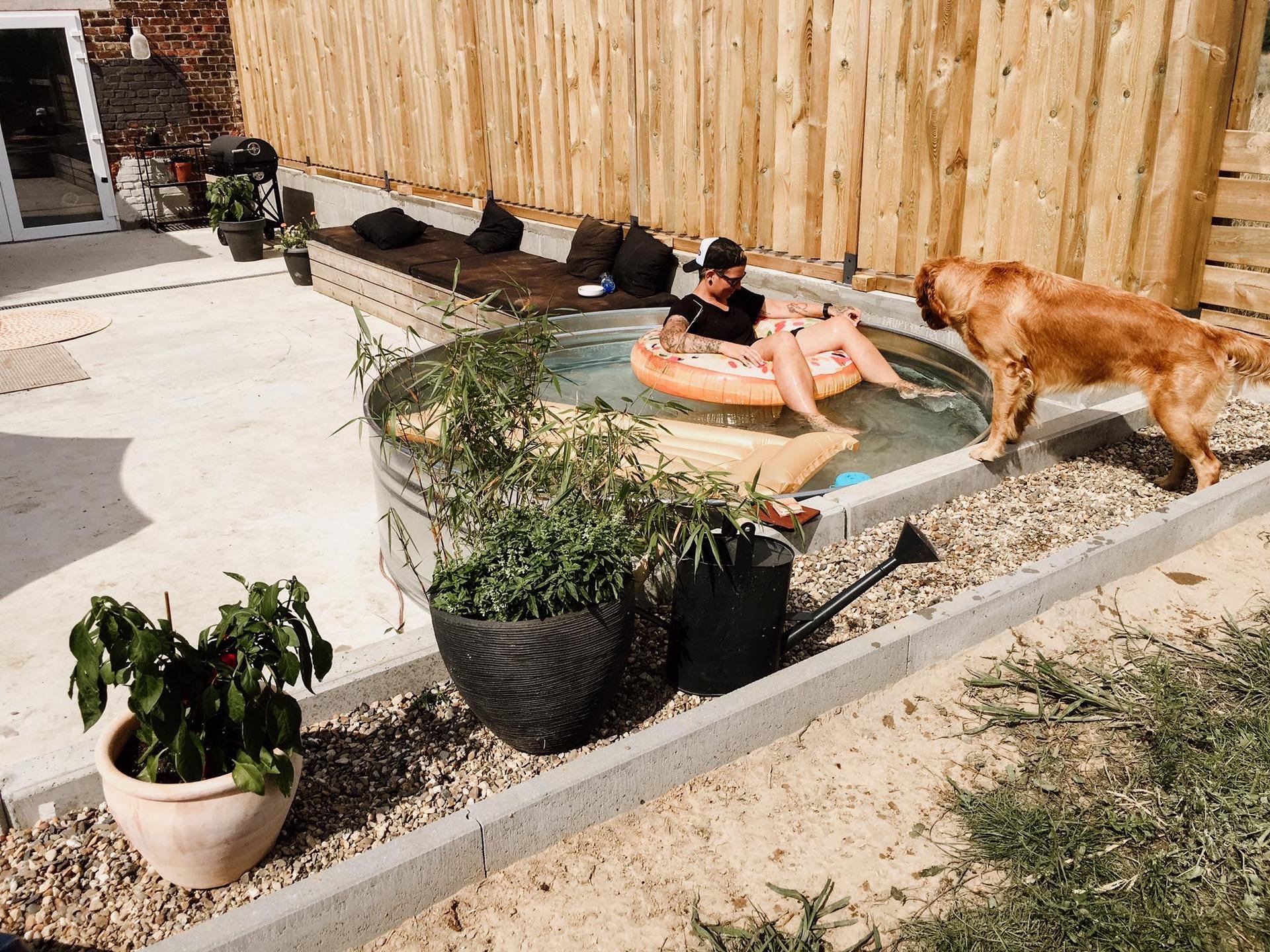 A woman relaxing in a Stock Tank pool with a floating lounge chair, and a golden retriever dog watching her, in a backyard enclosed by a wooden fence.
