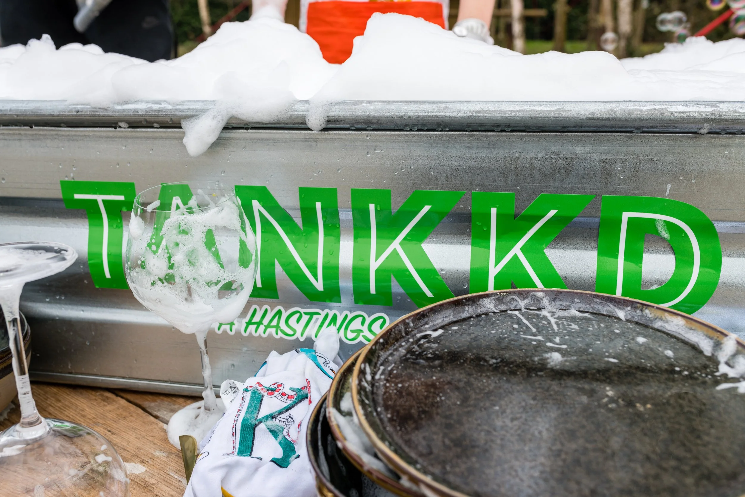 Cleaning foam on a boat with a green sign reading "TANK KIDD" and parts of glasses, cloth, and a tray or lid in foreground.