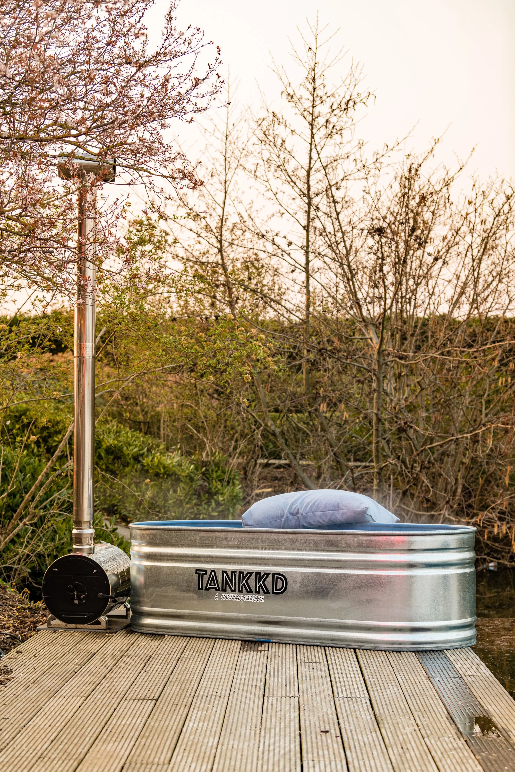 A metal bathtub labeled 'TANKKD' filled with water and steam, situated on a wooden dock near trees with budding leaves and pink blossoms, with a steam generator next to it.