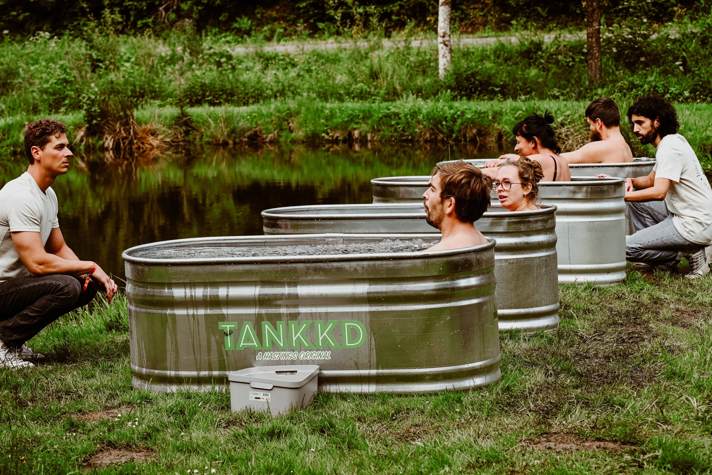 A group of people sitting in Stock tank ice baths filled with water near a lake, engaging in an outdoor activity on a grassy area surrounded by trees.