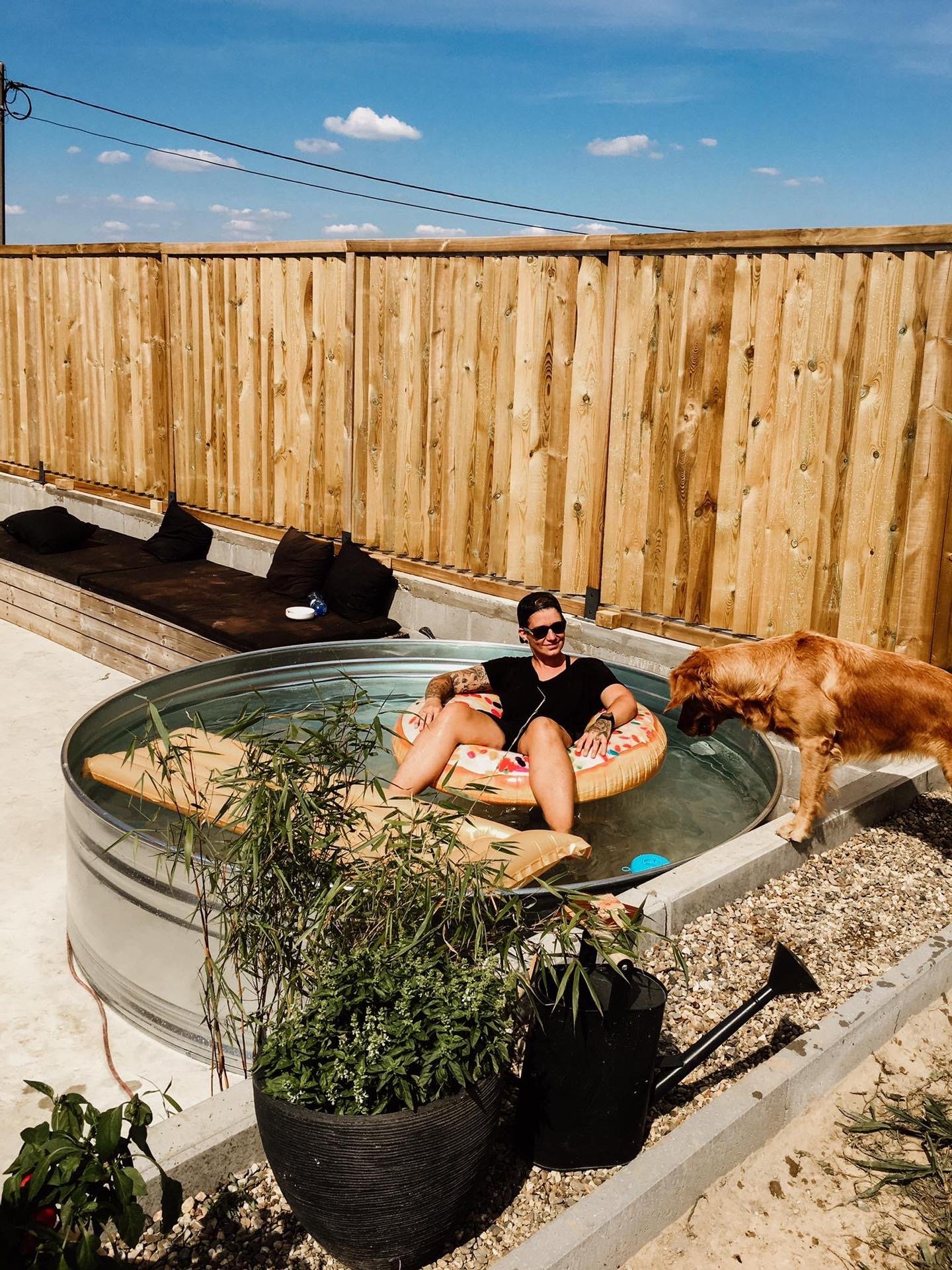 Woman relaxing in a small above-ground pool with a dog standing outside at the edge, in a backyard with a wooden fence, black cushions on a built-in bench, and potted plants.