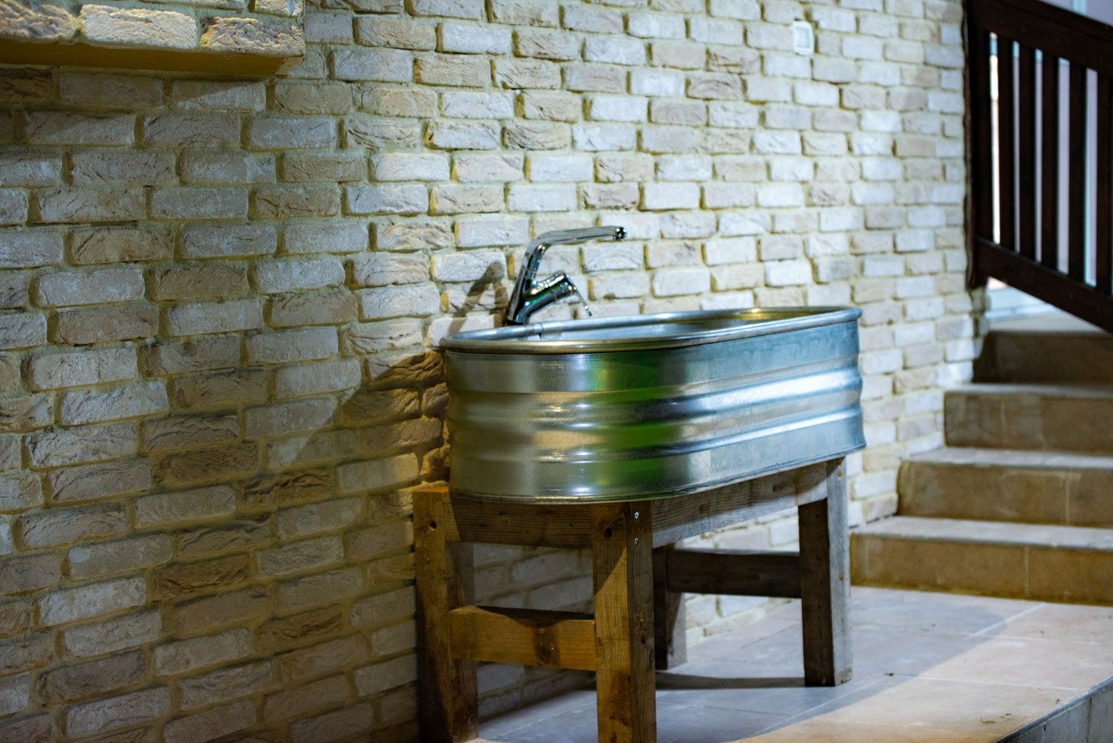 A rustic outdoor stone wall, with a metal wash tub on a wooden stand next to concrete steps and a staircase with a wooden railing.