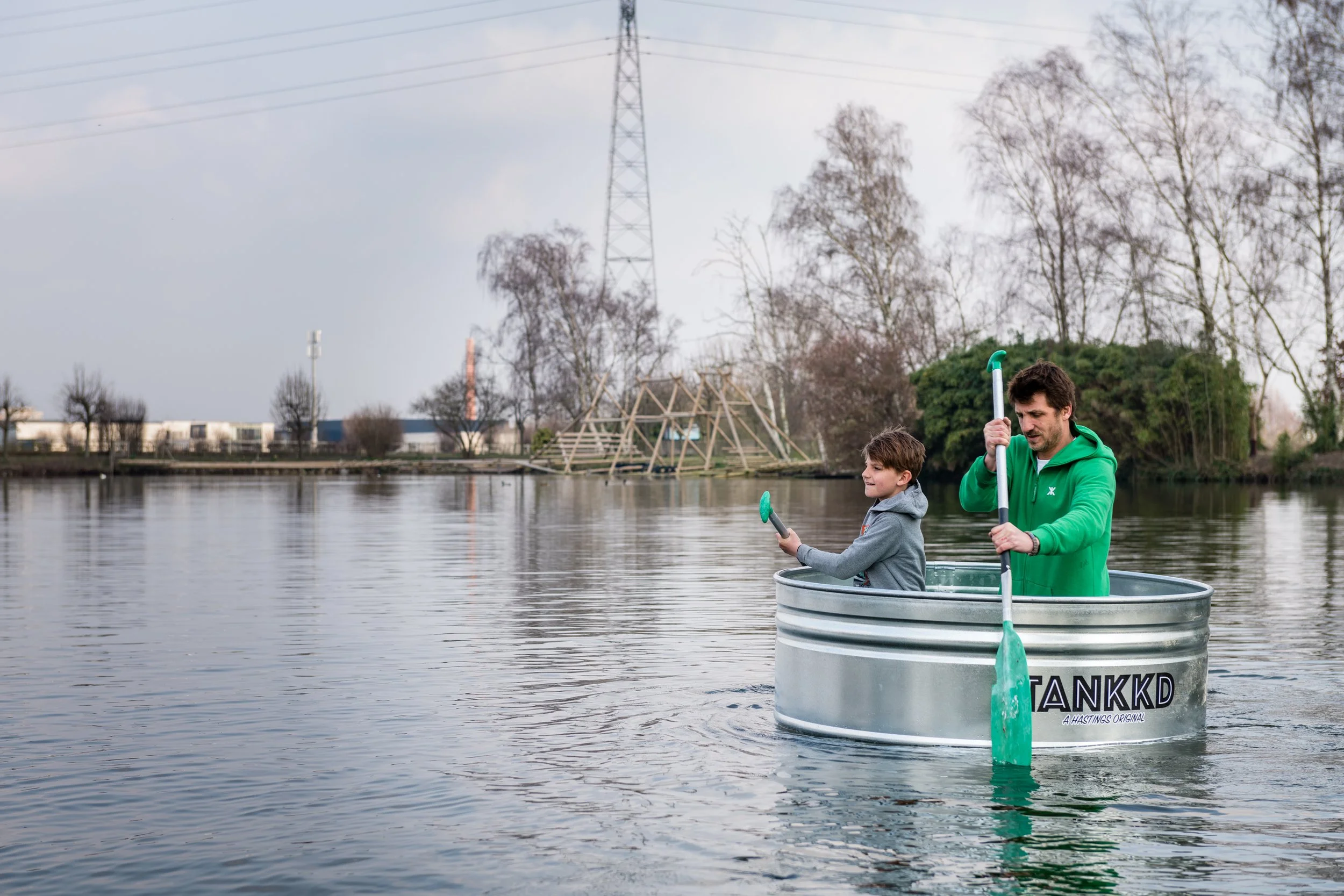 A man and boy in a metal tub on a calm lake, using paddles and a fishing rod, with trees and a wooden structure in the background.