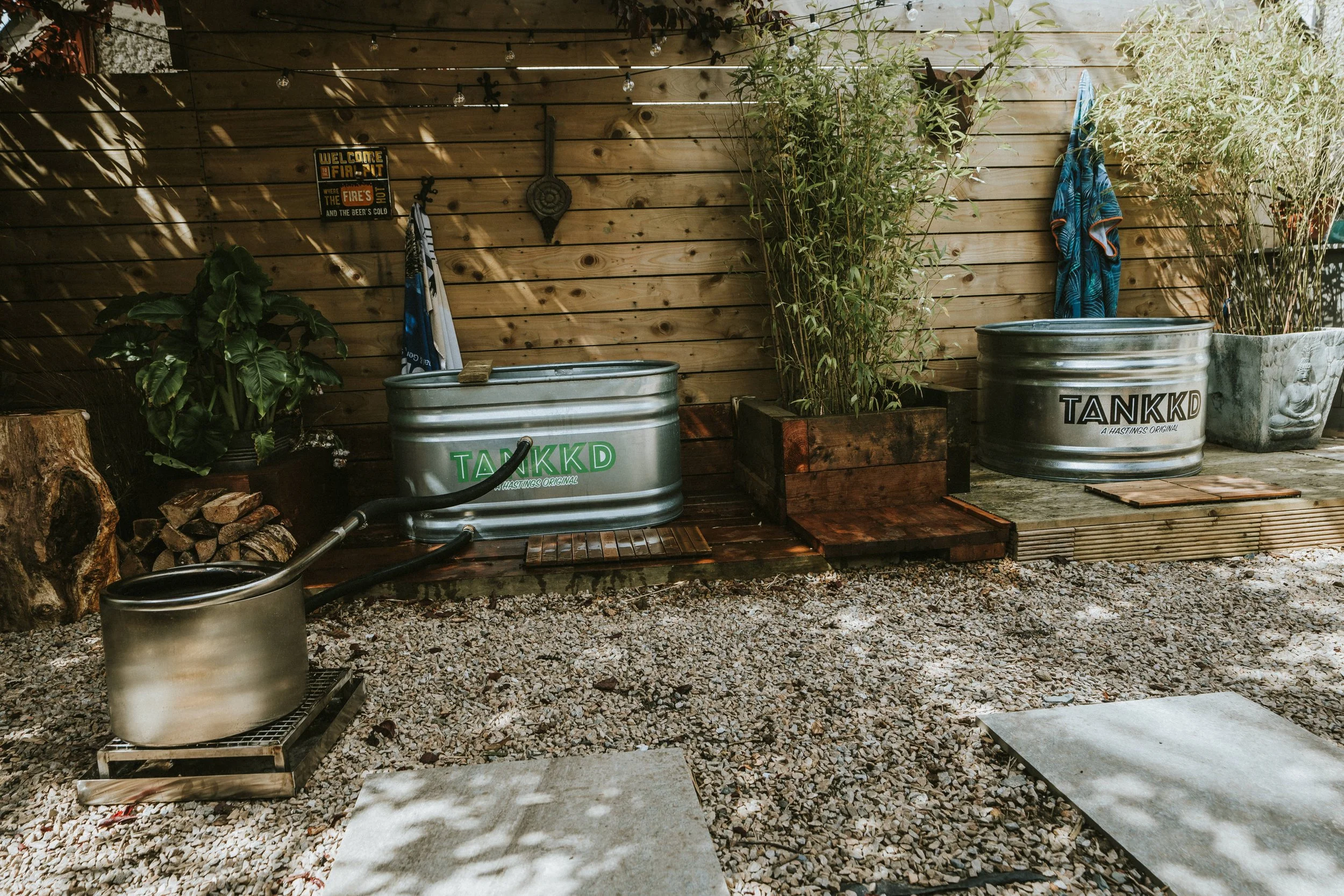 Outdoor backyard space with wood privacy fence, Stock tank pool , hot tub and ice bath firewood, gravel ground, and a small grill with a pot on top.