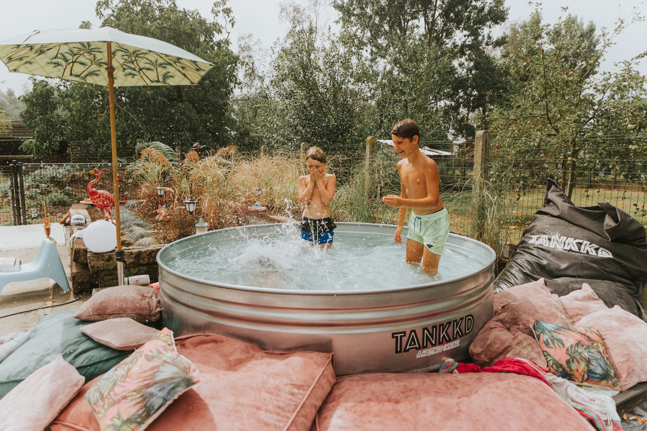 Two children playing in a small above-ground pool with a garden and trees in the background. The girl is covering her face while the boy is smiling. The pool is surrounded by cushions and pillows on outdoor furniture.