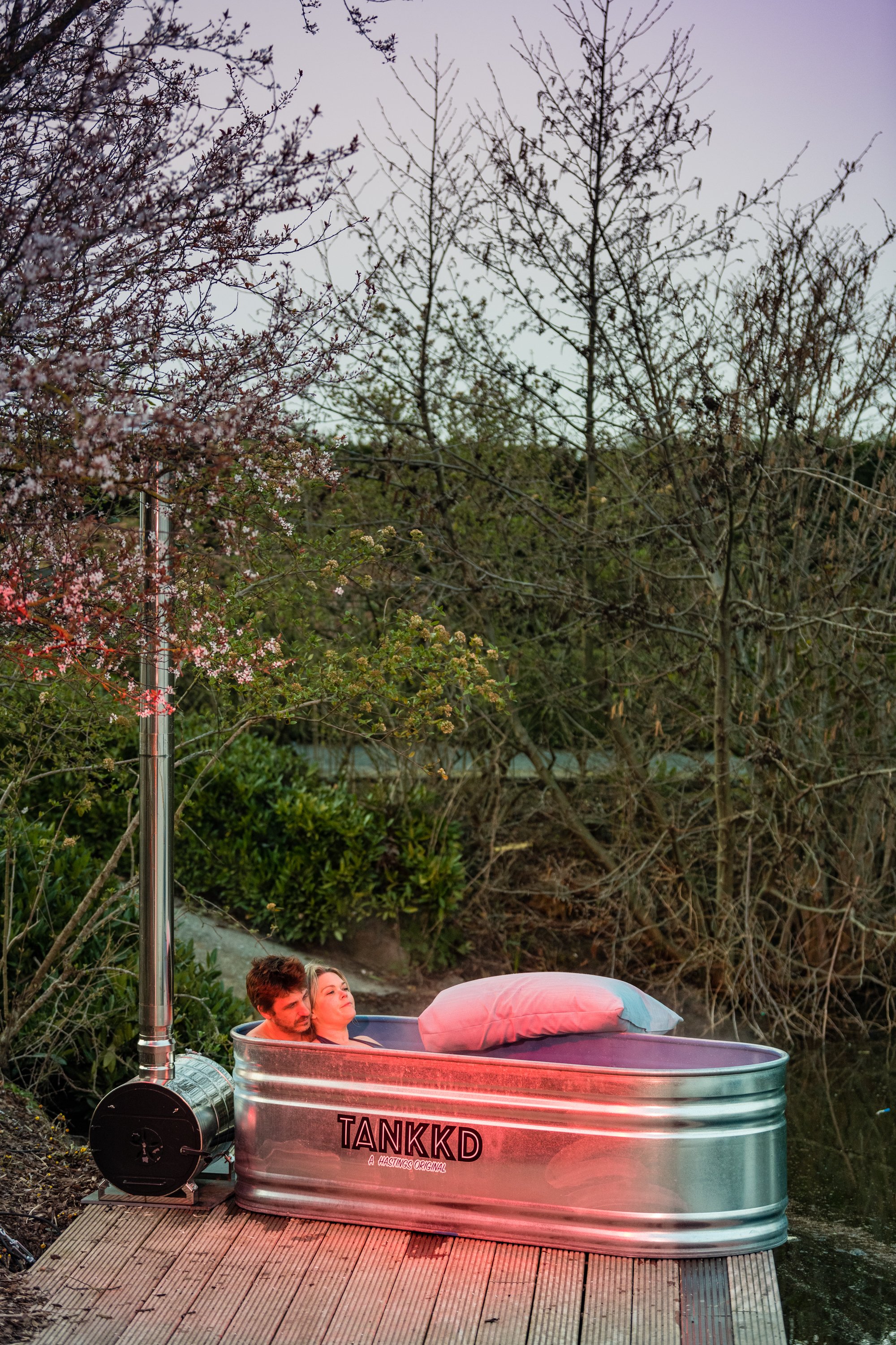 A couple relaxing in a Stock tank bathtub made from a metal water tank labeled 'TANKKD' outdoors on a wooden deck surrounded by trees and bushes.