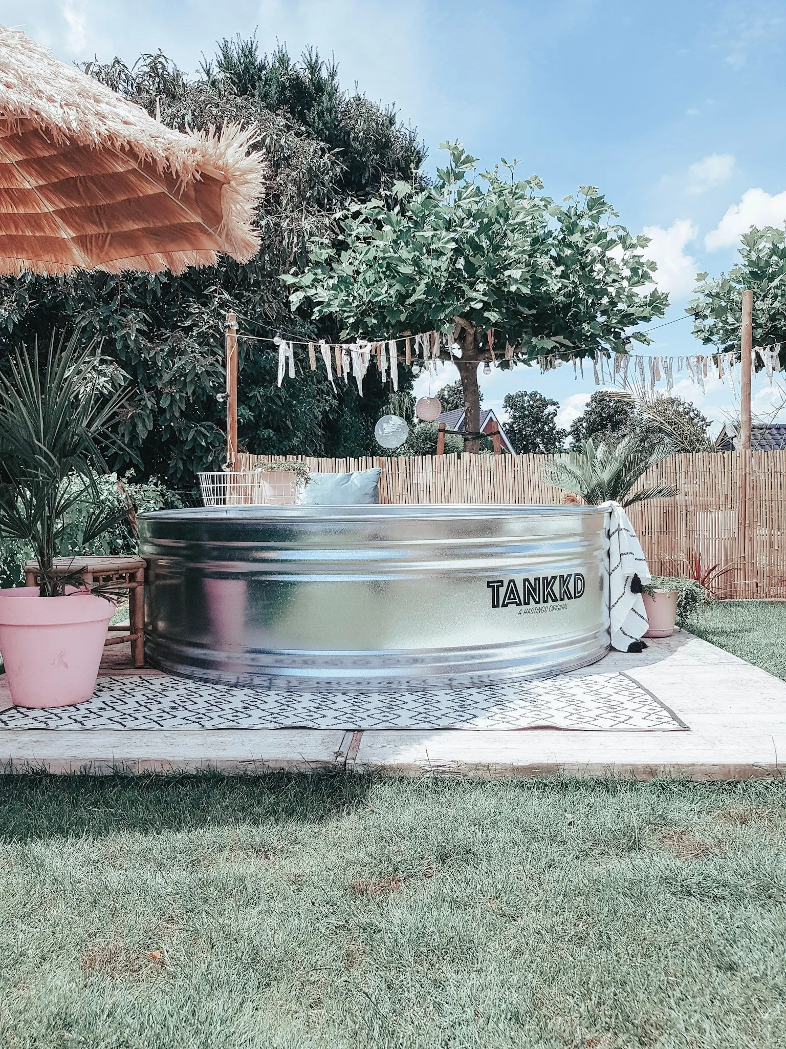 A backyard with a Stock tank pool , pink potted plants, a bamboo privacy fence, string lights, and a large tree under a blue sky with some clouds.