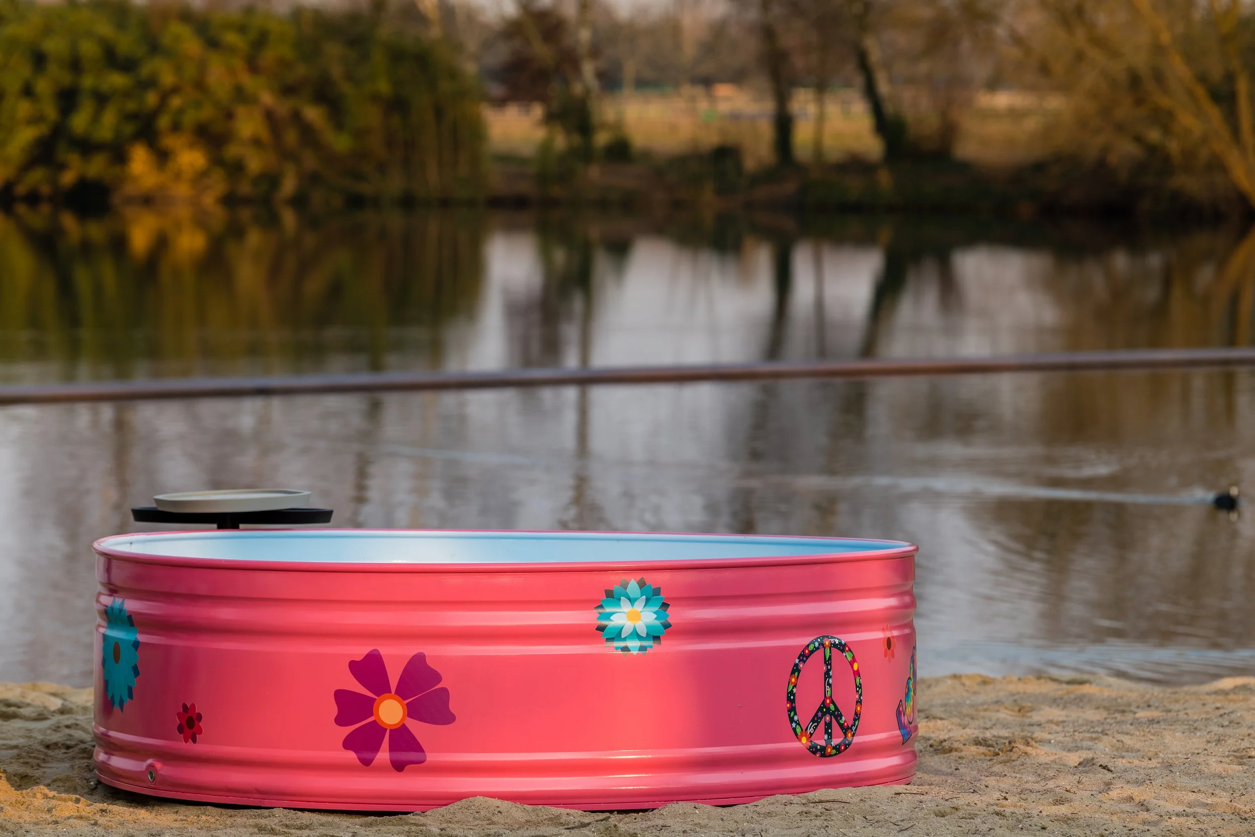 A pink metal container decorated with flowers, peace signs, and heart patterns on a sandy beach near a calm body of water, with trees and a distant shoreline in the background during sunset.