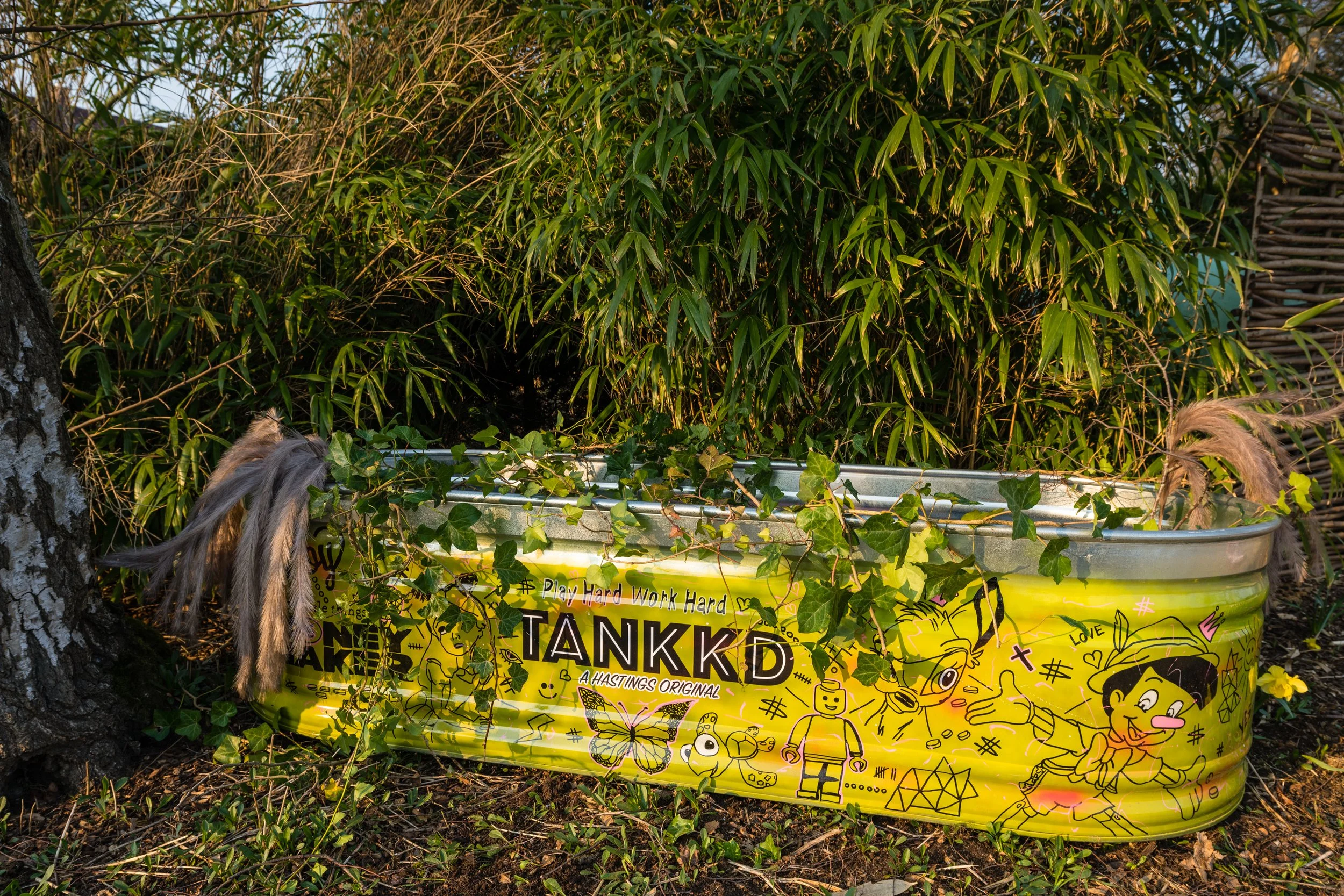 A yellow metal container with graffiti and illustrations, placed outdoors near green plants and a tree trunk, with ivy climbing on it.