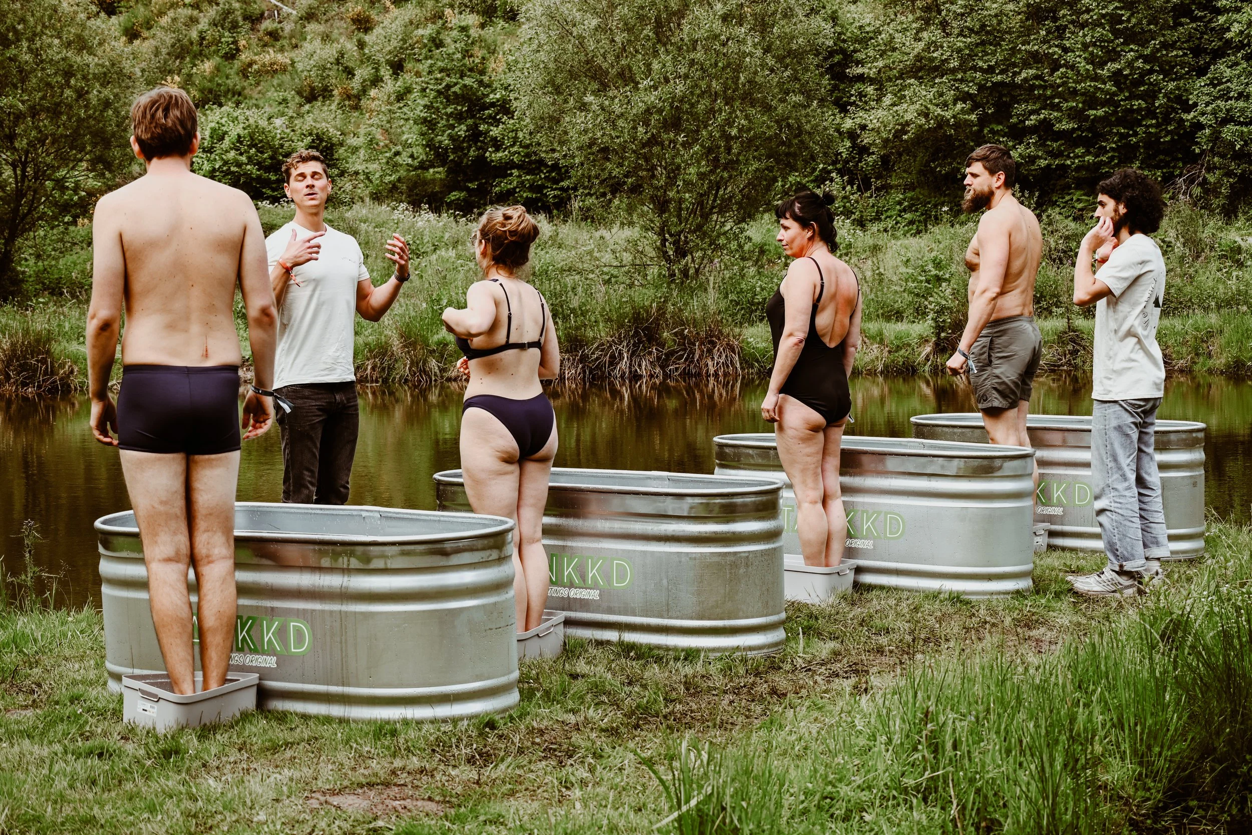 A group of people standing in water-filled Stock tank pool  outdoors near a pond, participating in a discussion or ceremony surrounded by lush green trees and grass.