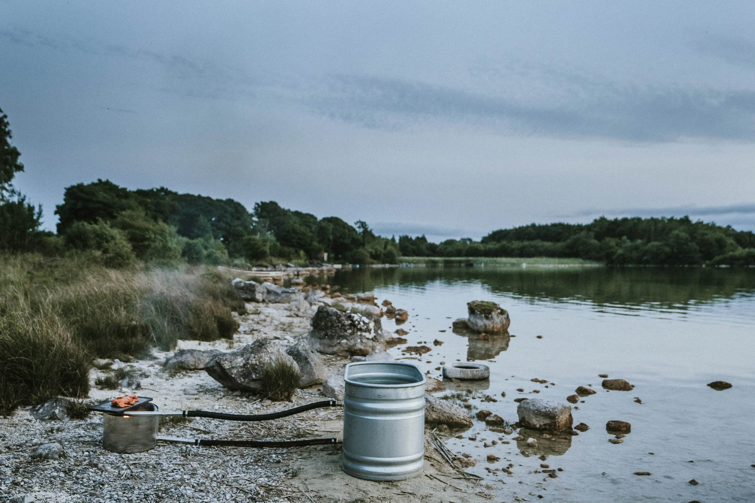 A serene lakeside scene with a metal barrel, a hose, and a small pot on the rocky shore, lush green trees in the background, calm water, and an overcast sky.