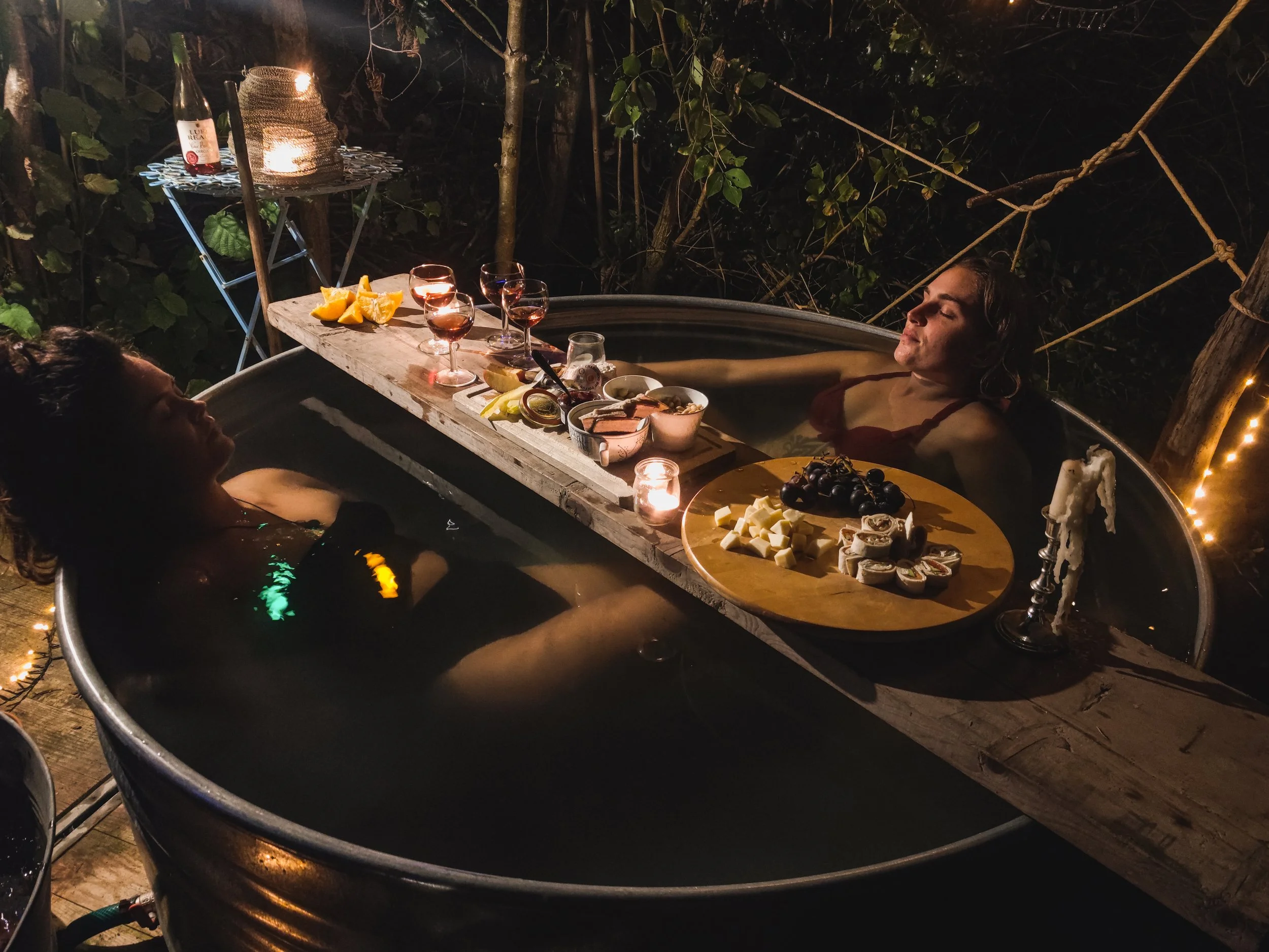 Two women relaxing in a hot tub at night with a wooden table between them holding snacks and drinks, surrounded by string lights and outdoor greenery.