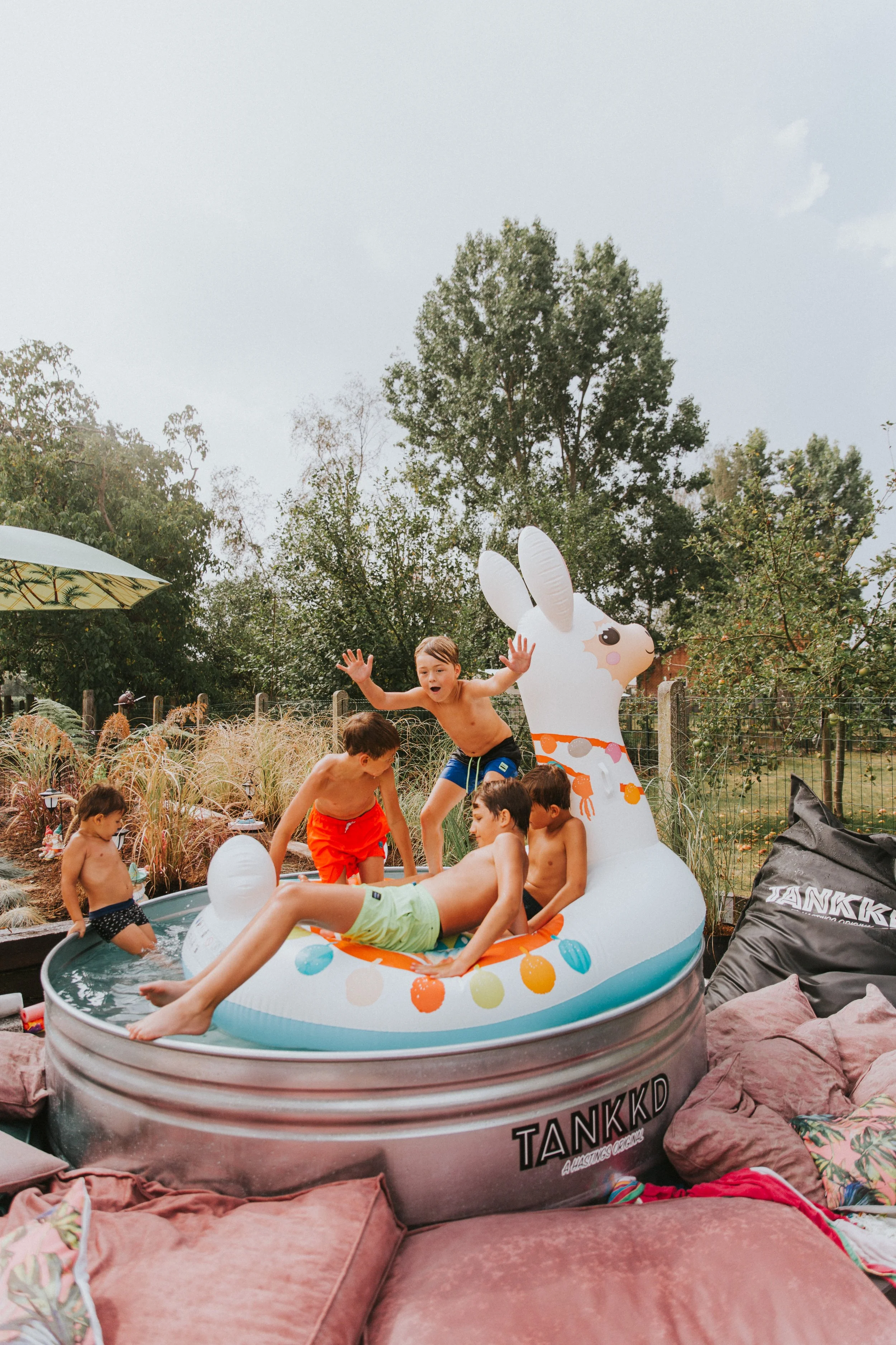 Five children playing in a Stock tank pool with a llama design in a backyard. One child is lying on the pool, and four others are jumping or standing around it. The backyard has trees, grass, and outdoor furniture.