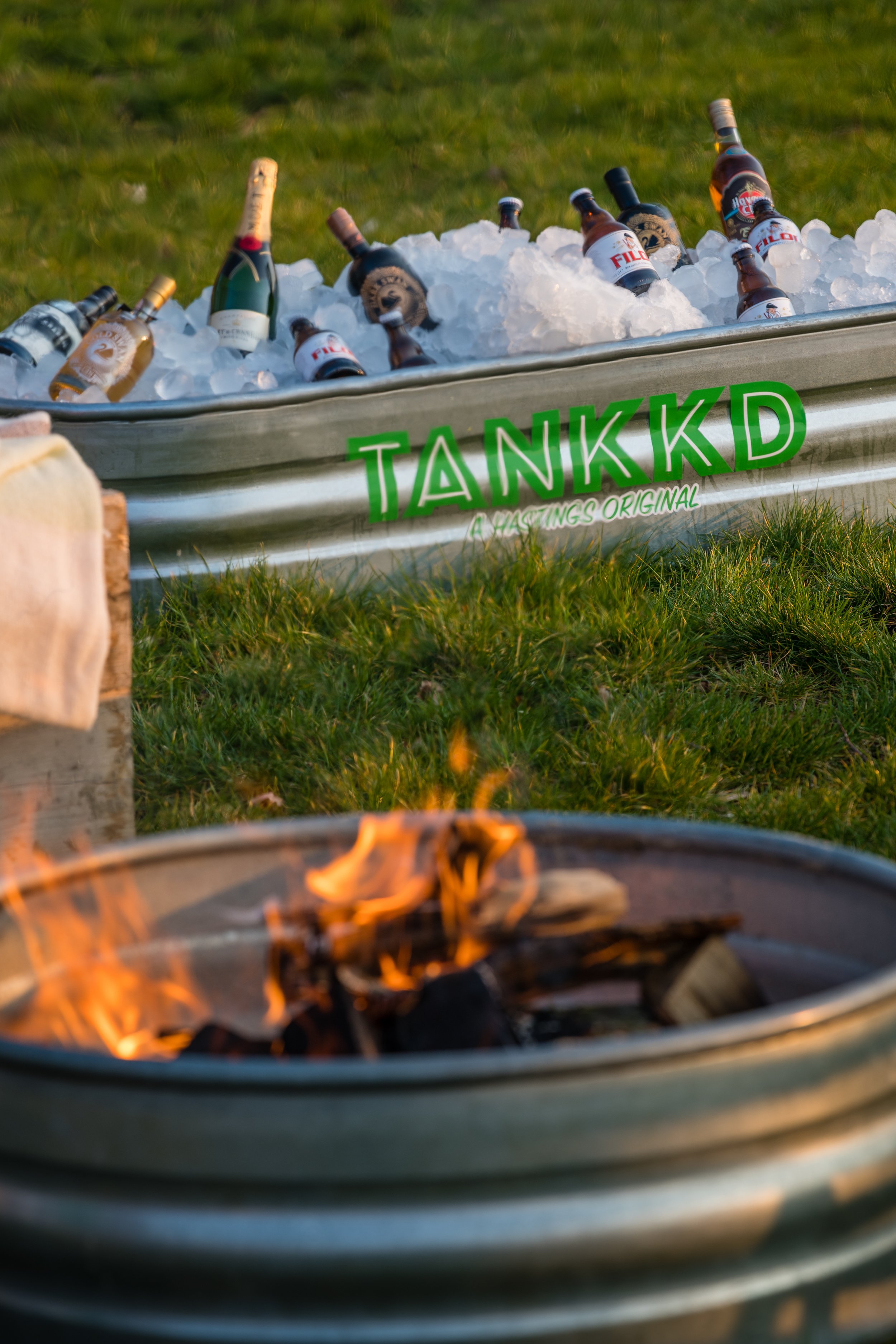 A Stock Tank filled with ice and bottles of beer, labeled 'TANKKD', on grass, with a campfire in the foreground.
