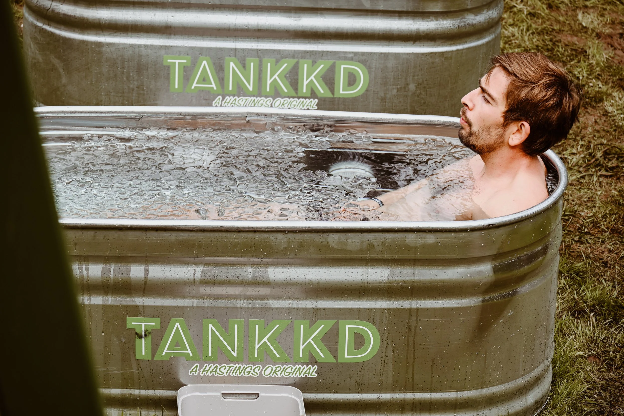 A man in a ice bath outdoors, with a sign on the tub that reads 'TANKKD: A Hastings Original'.
