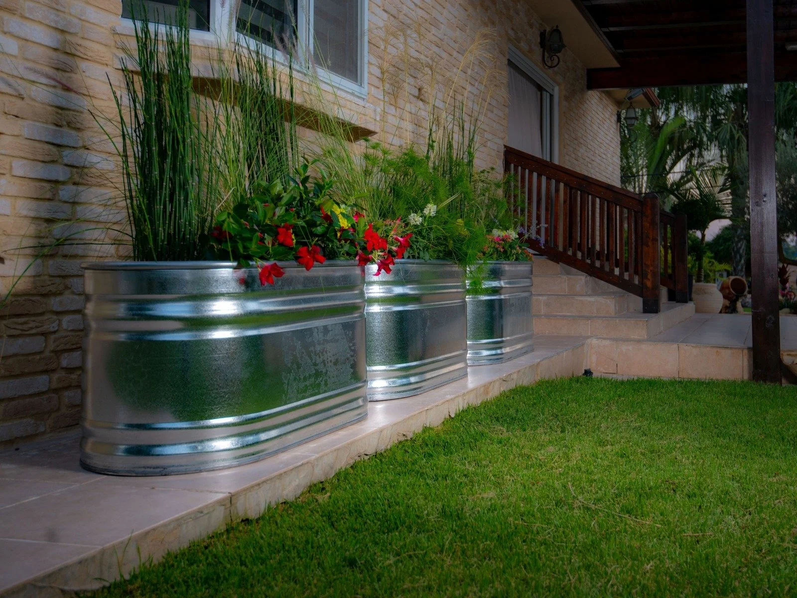 Stock Tank garden planters with colorful flowers and tall grass on a patio next to a brick house with stairs leading up to a porch.