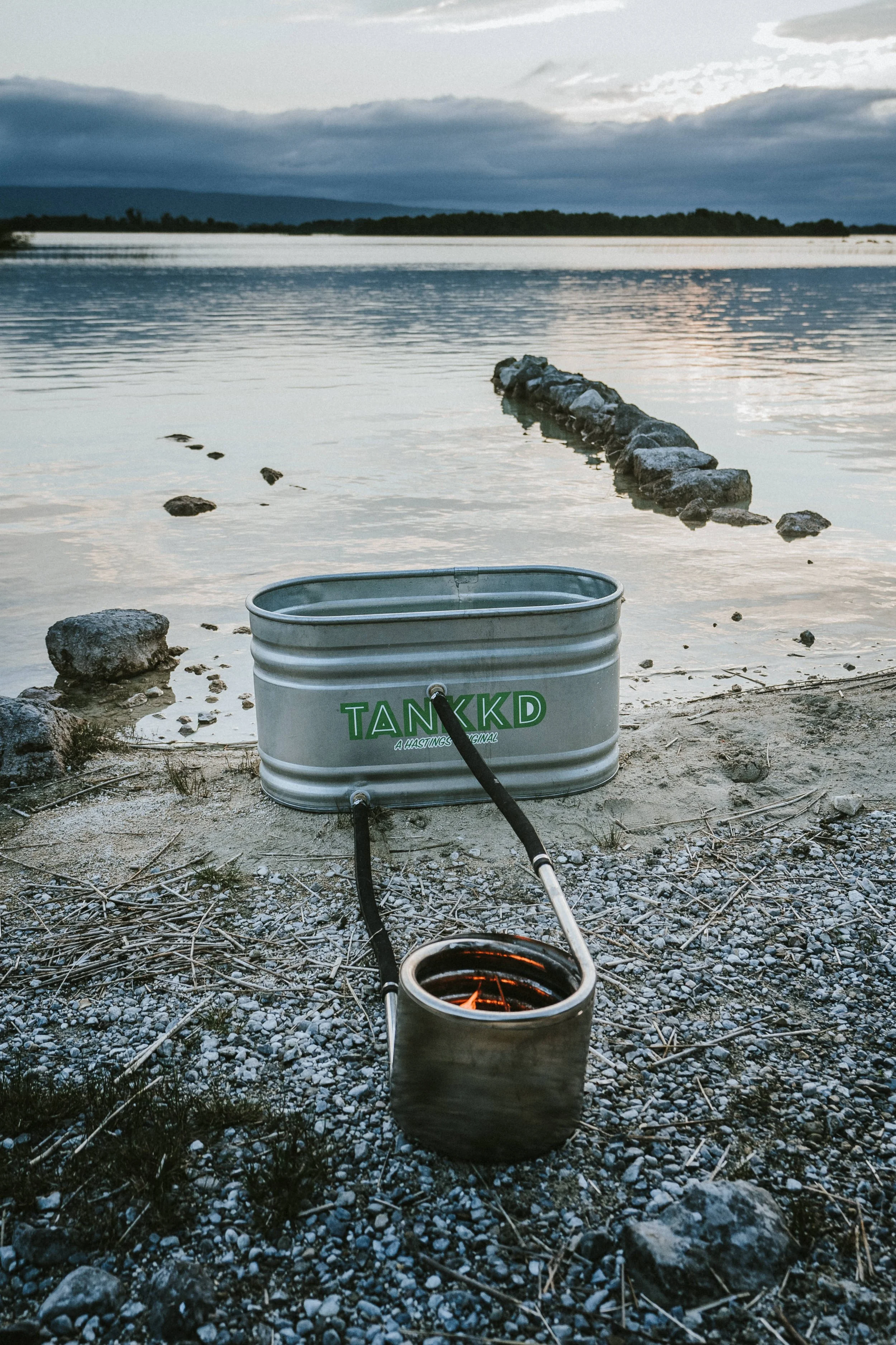 A metal water container labeled 'TANKKD' with hoses attached, placed on rocky shores near a calm lake with a stone jetty extending into the water, during a cloudy sunset.