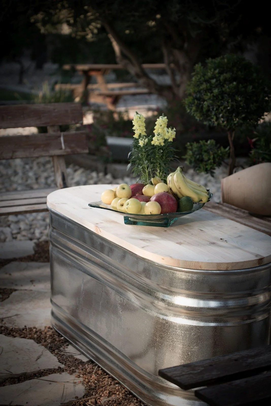 A glass dish filled with various fruits including bananas, apples, and limes, with a yellow flower bouquet, placed on a wooden table outdoors.