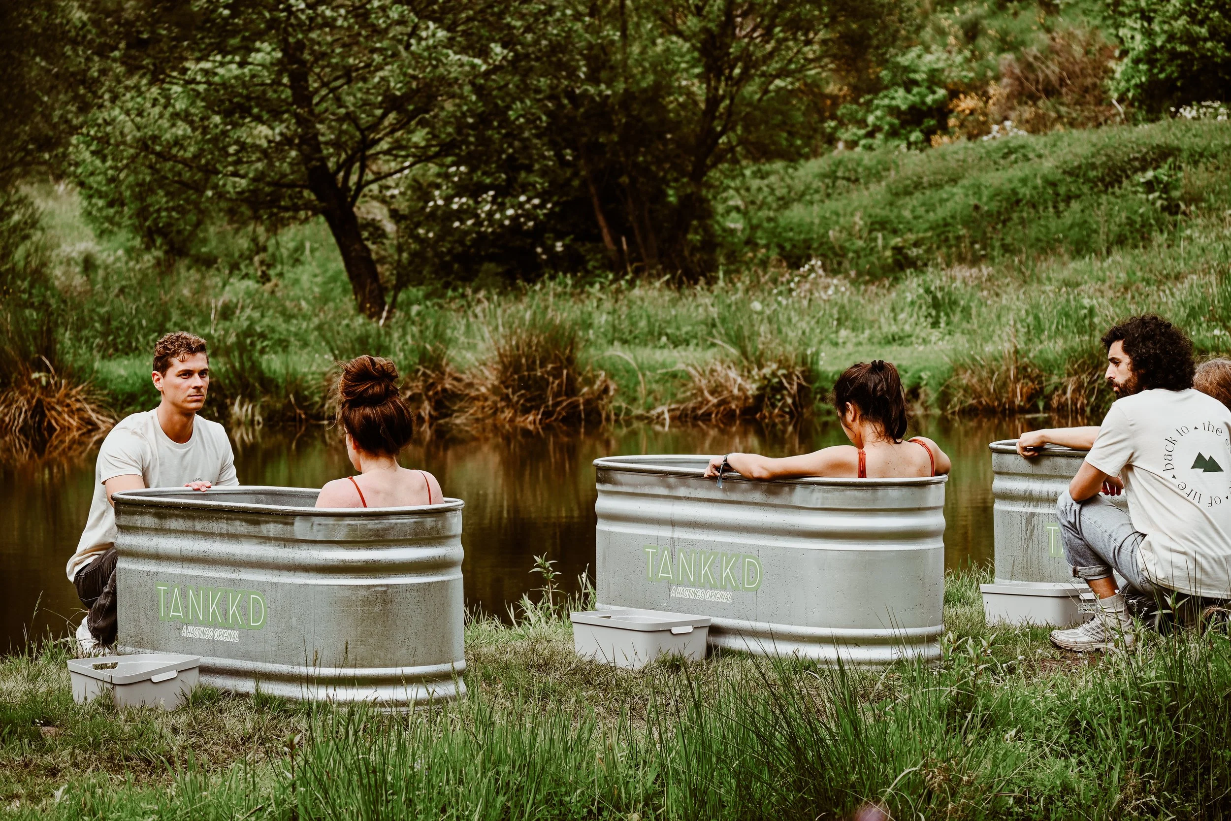 Four people sitting in ice baths by a river, relaxing outdoors with greenery in the background.