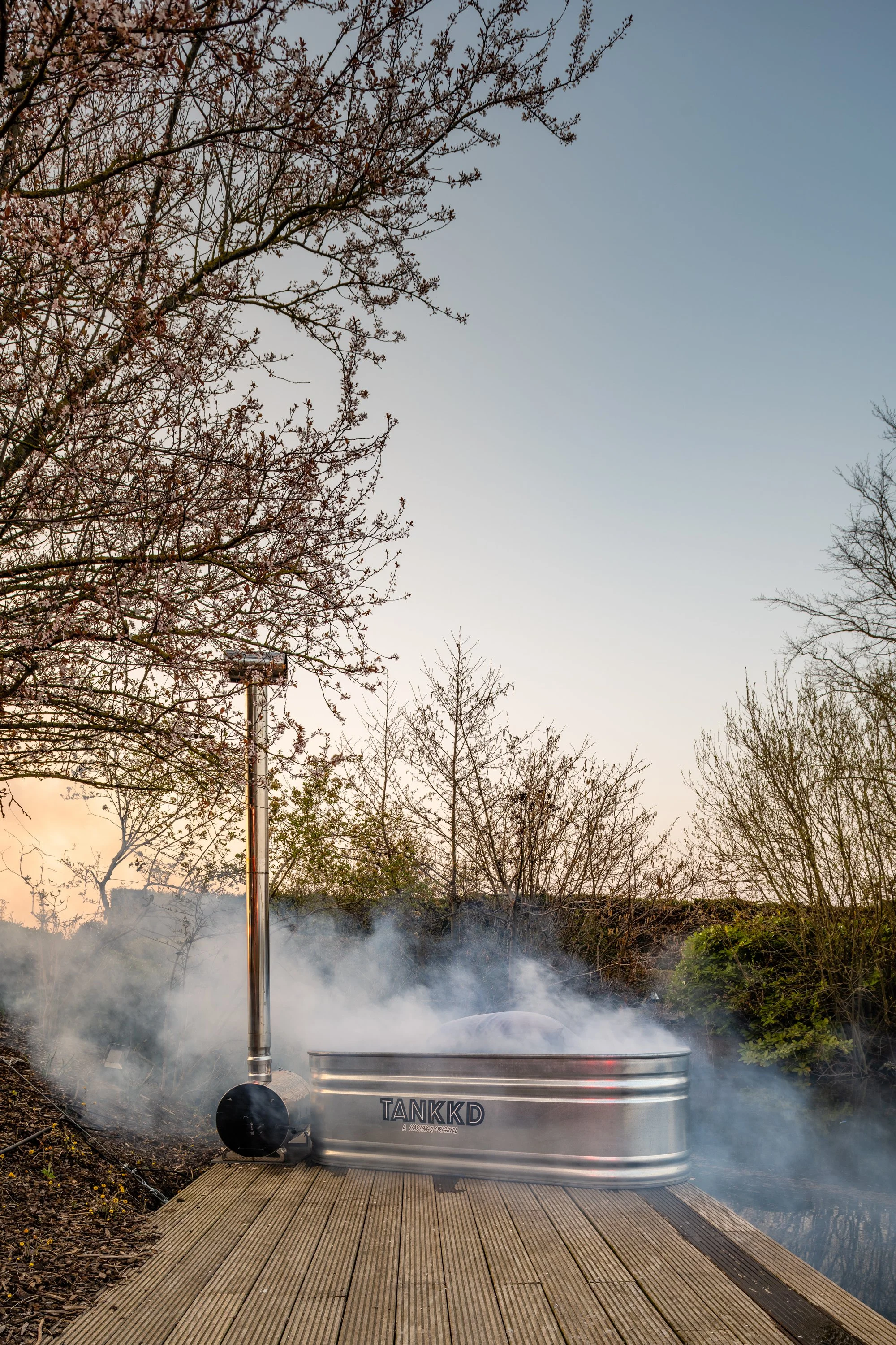 An outdoor hot tub emitting steam on a wooden deck, surrounded by trees and plants during sunset.