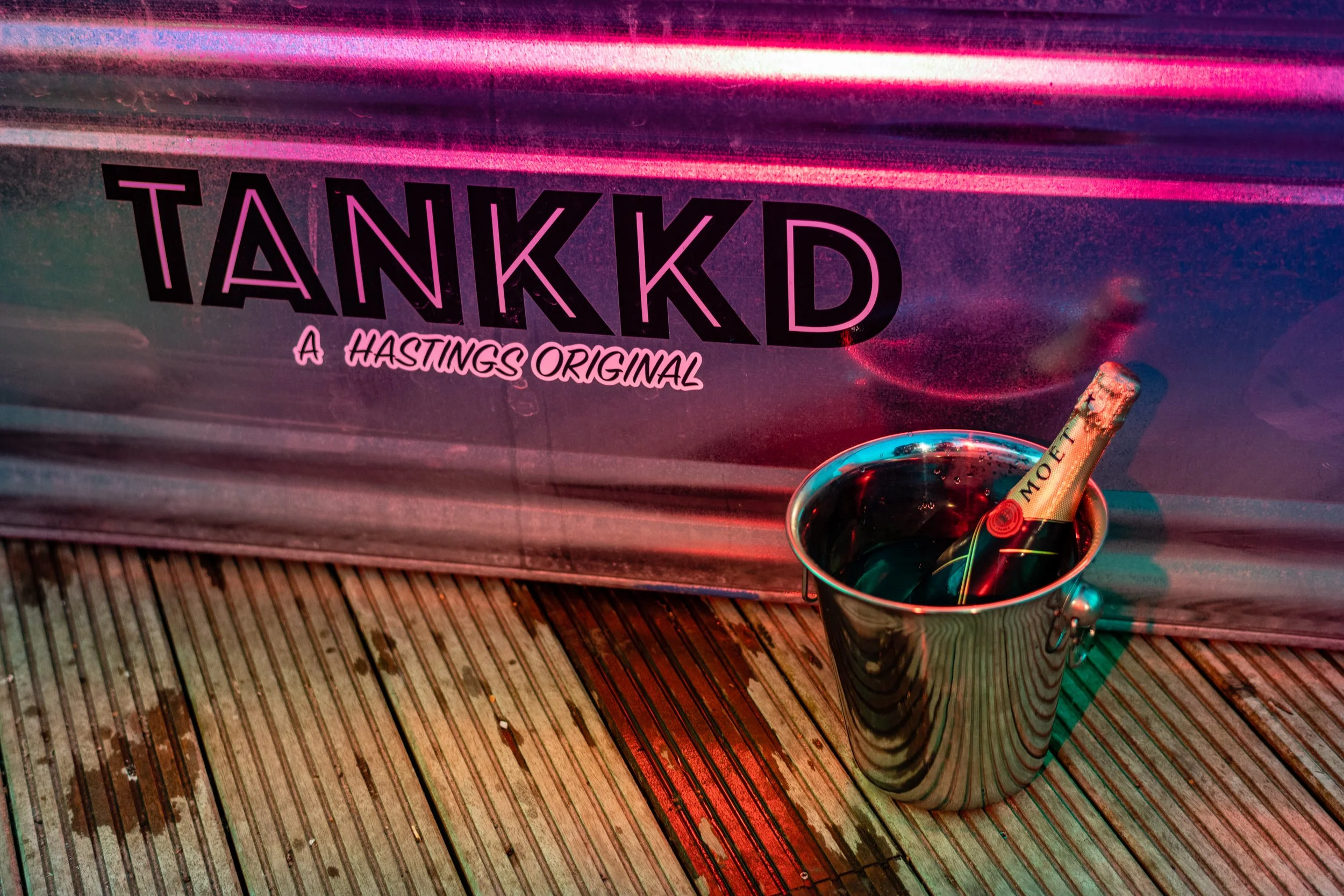 A metal bucket with a bottle of Moët champagne inside, placed on a wooden deck. Background has a colorful sign with the text "TANKKD, A HASTINGS ORIGINAL" in neon pink and black.