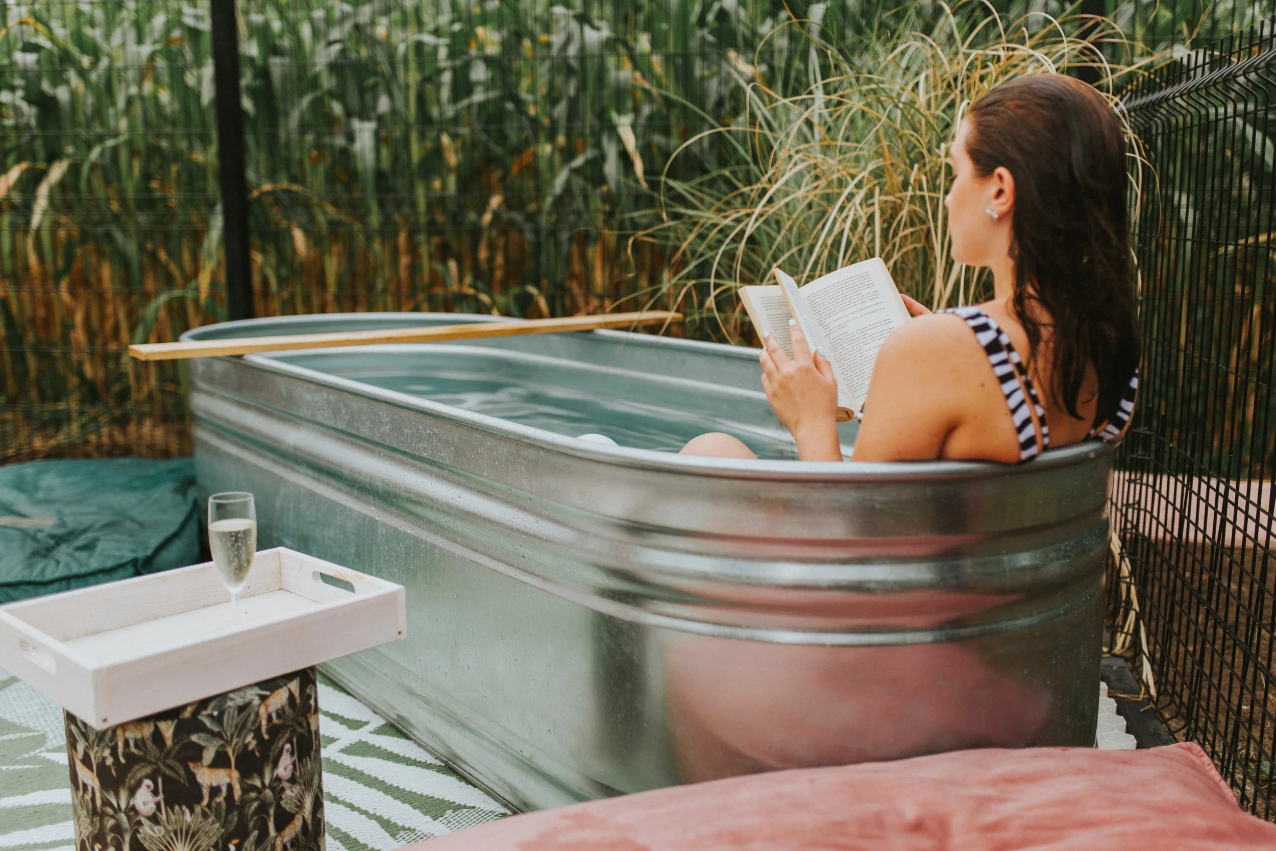 Woman sitting in a Stock tank pool  filled with water, reading a book, outdoors surrounded by tall green plants and grass, with a glass of champagne on a tray nearby.