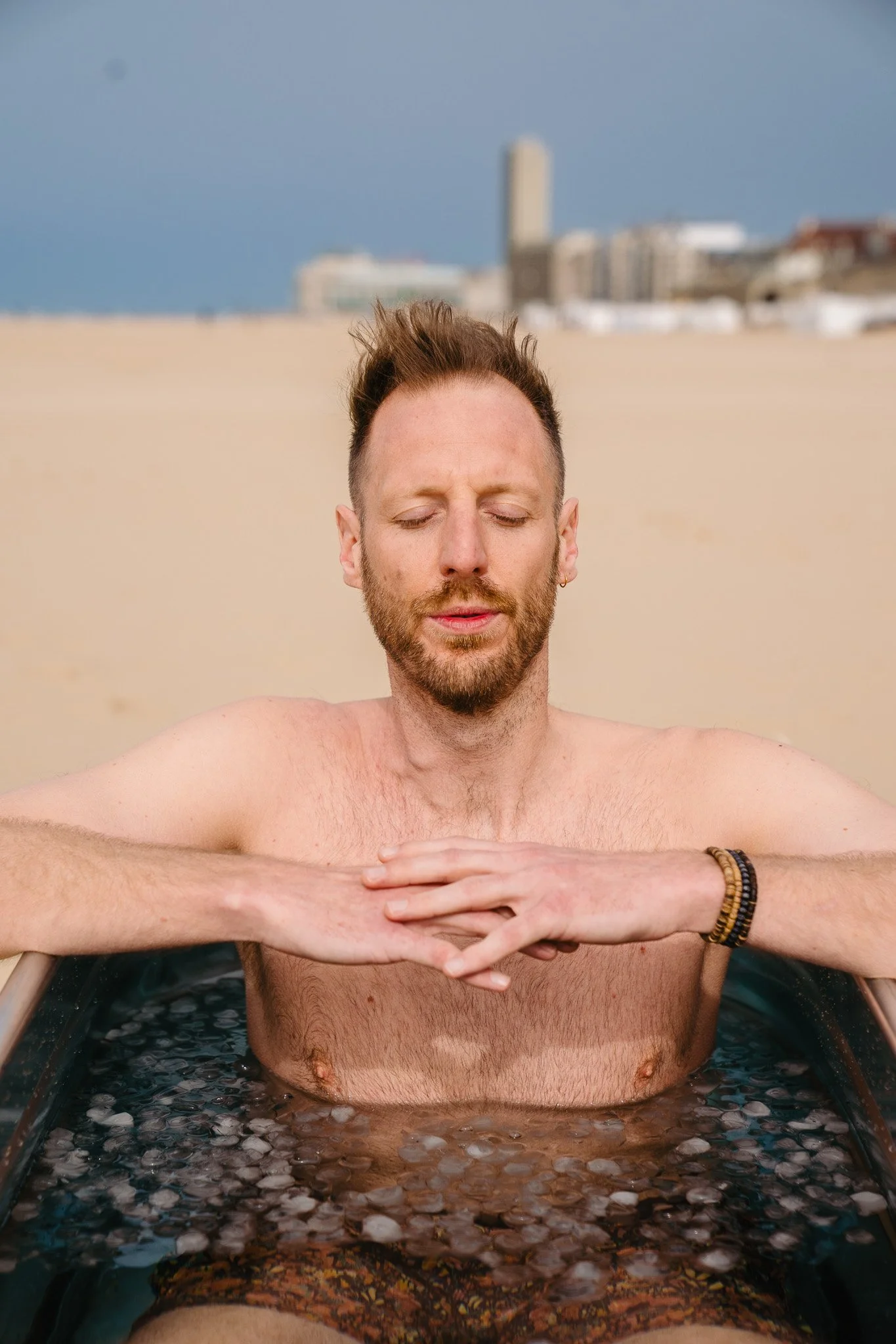 A man with closed eyes and beard relaxing in an outdoor hot tub on a beach, with a city skyline in the background.