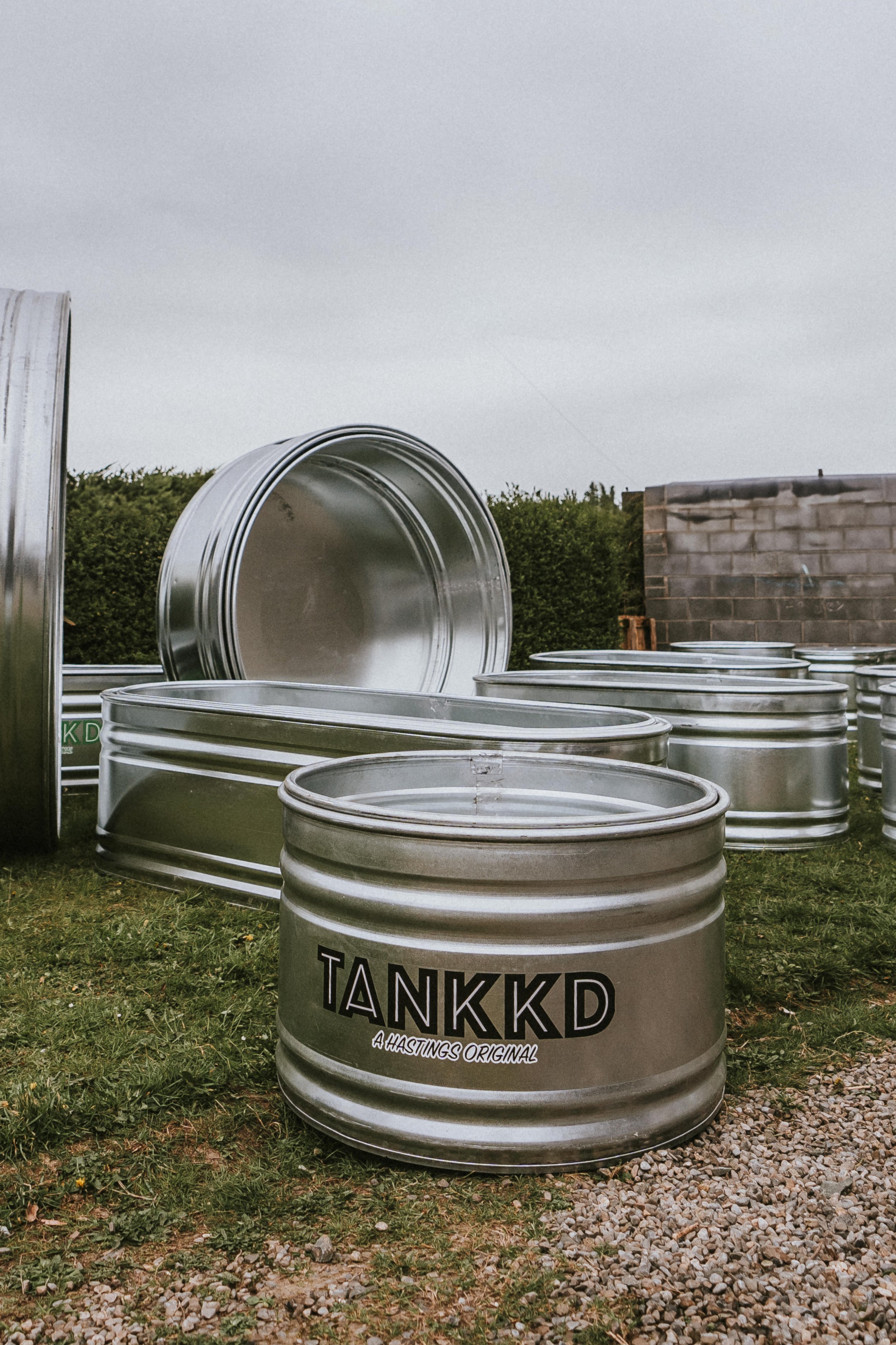 Multiple stacked stock tanks outdoors on grass and gravel, some with brand labels, with trees, bushes, and a cloudy sky in the background.