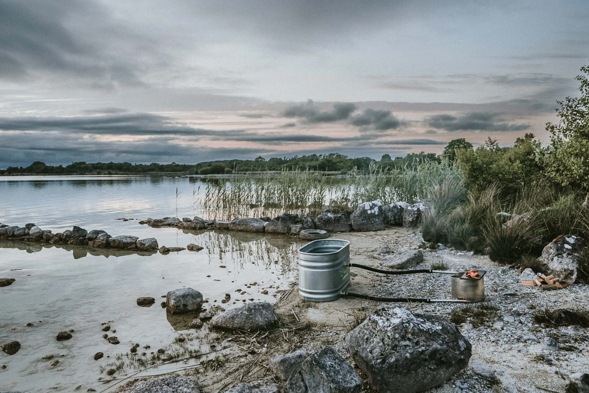 A serene lakeside scene with a cloudy sky, calm water, reeds in the background, and a wood coil burner connected to a 4ft stock tank on it on the rocky shore.