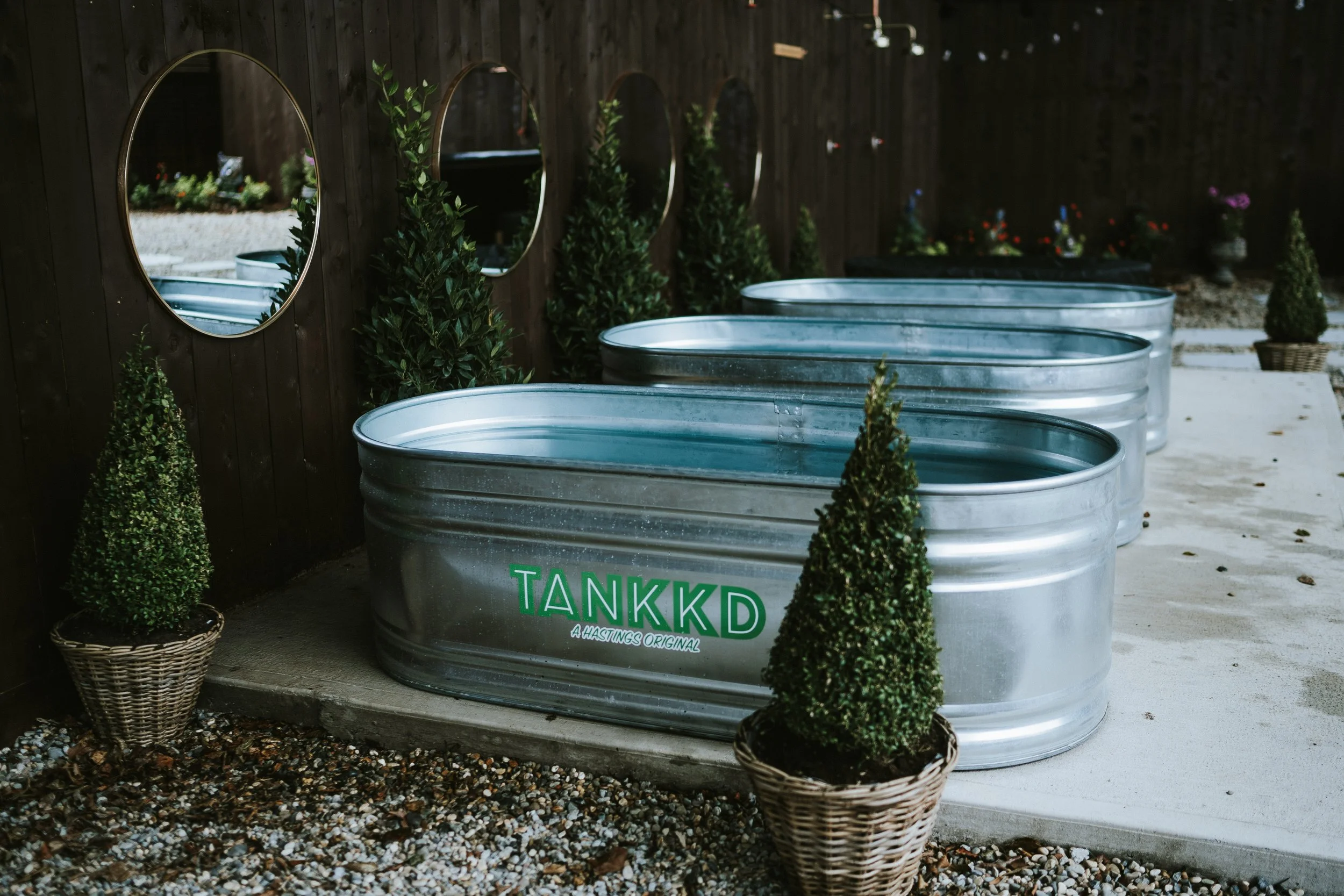 Three galvanized metal wading pools with green 'TANKKD' logo, lined up on concrete patio, surrounded by potted and planted greenery, against a wooden fence with oval mirrors.