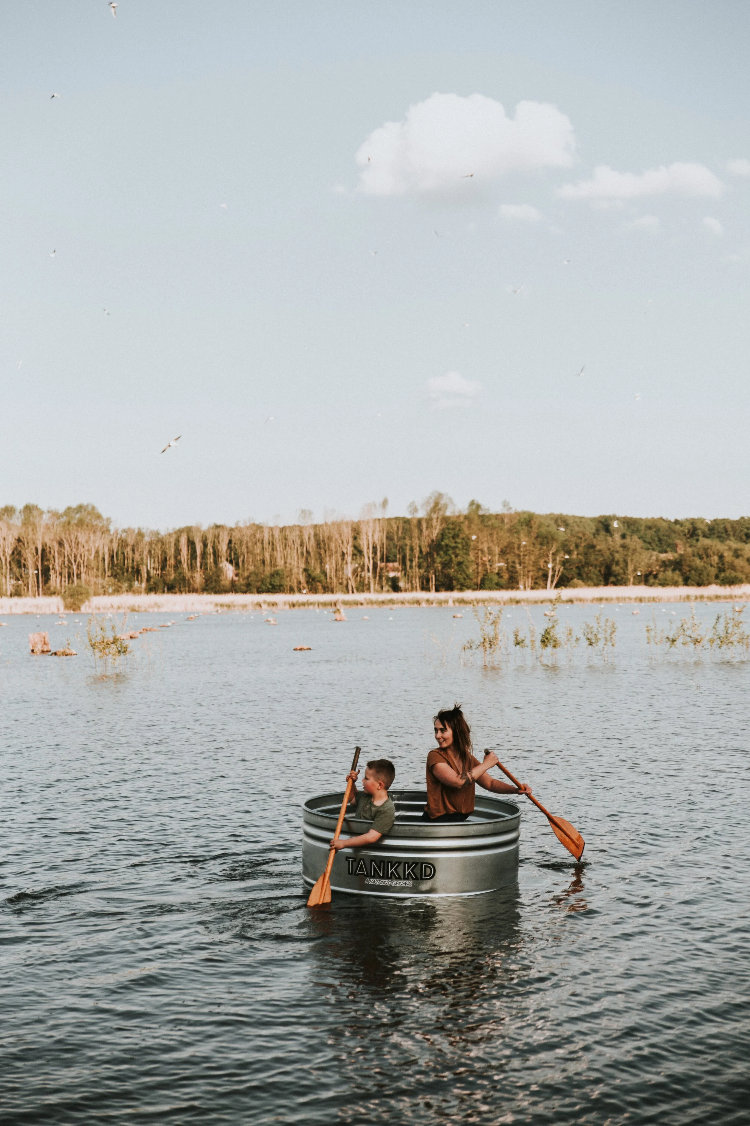 A woman and a young boy are sitting in a Stock tank on a body of water, each with an oar, as they enjoy paddling on a sunny day with a clear sky and some clouds.