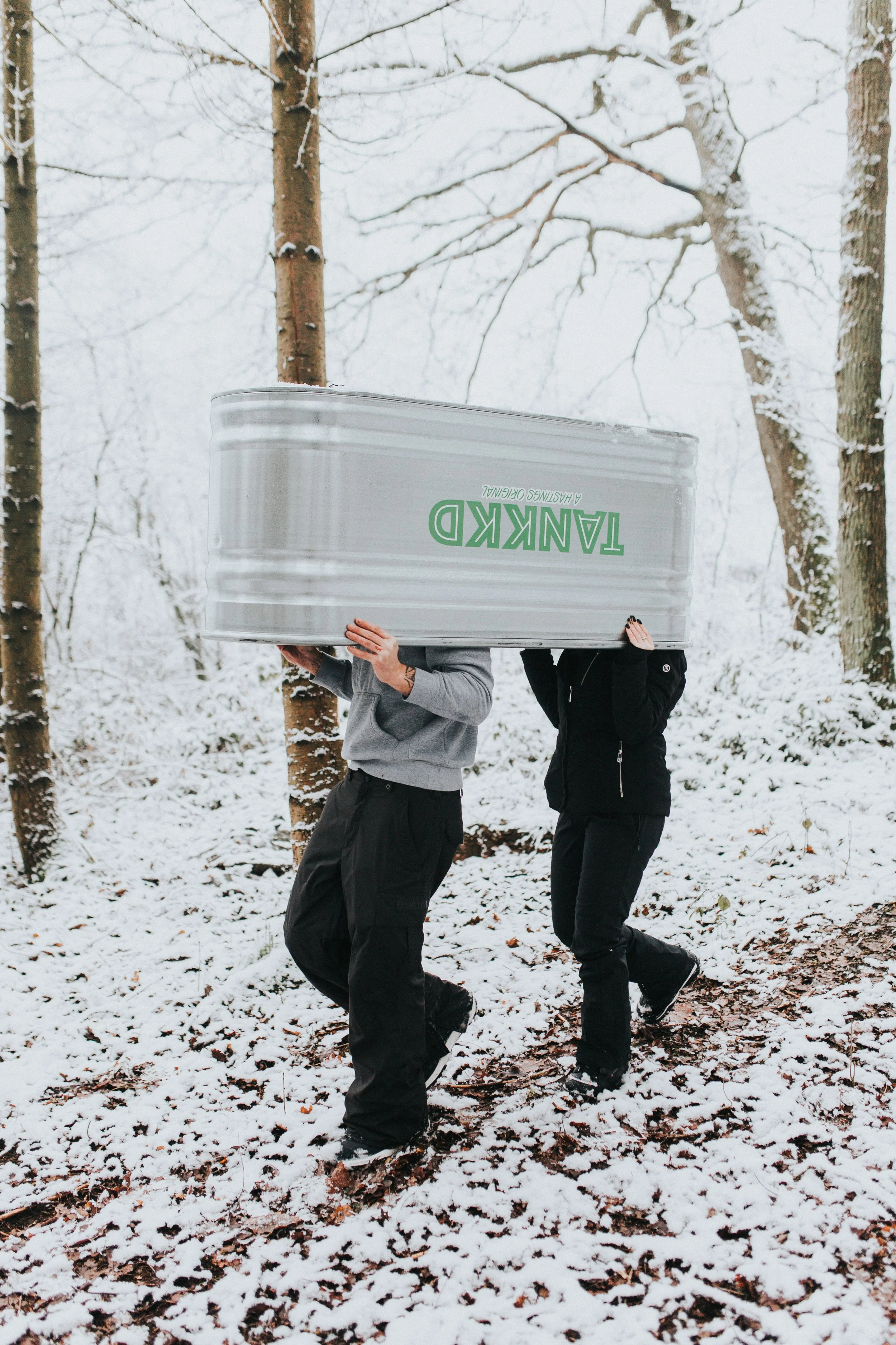Two people hiking through a snow-covered forest, carrying a largeStock tank labeled 'TANKED' with green text, with trees and snow in the background.