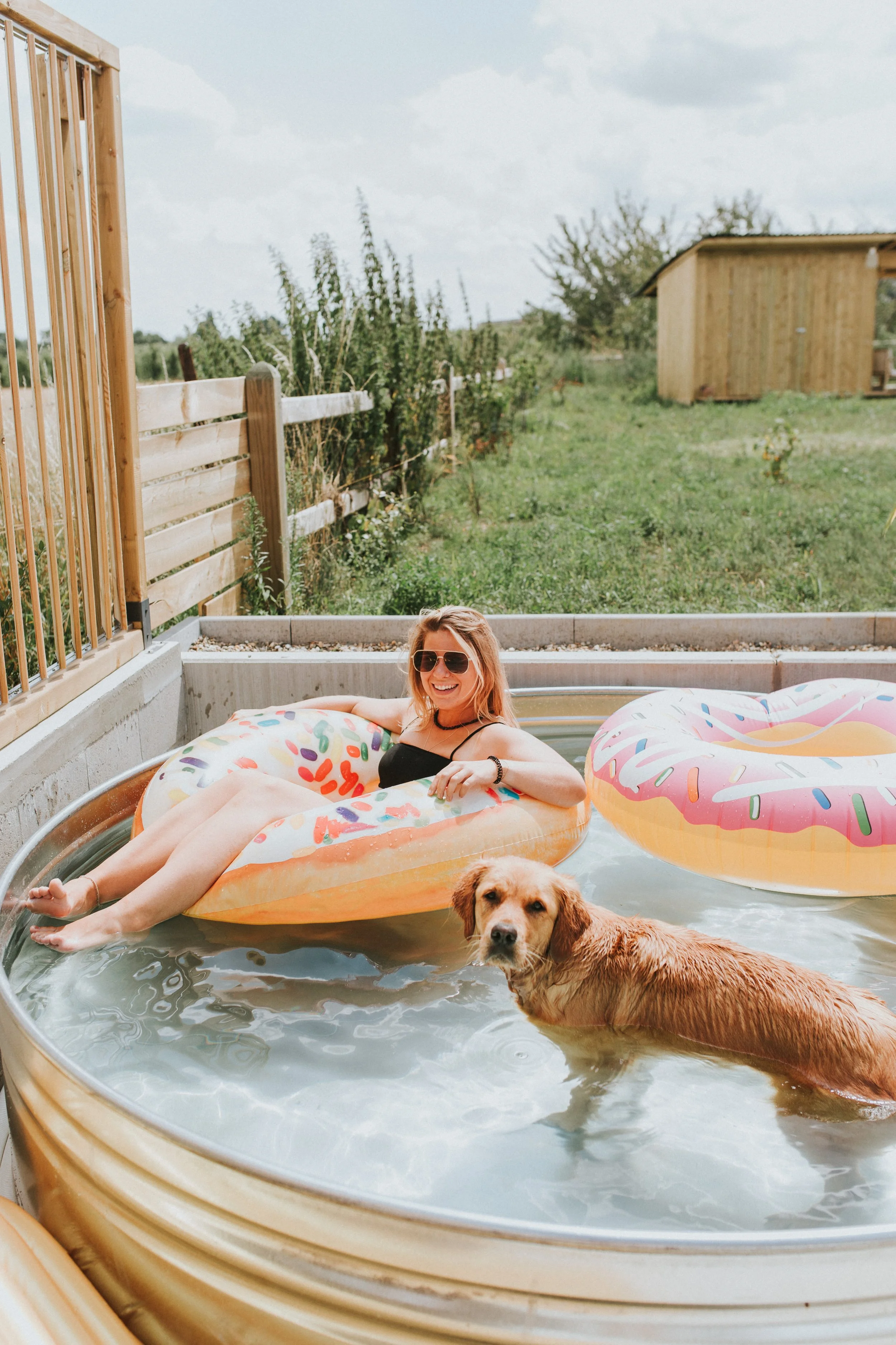A woman relaxing on a float in a small above-ground Stock tank pool with a dog in the water, outdoors on a sunny day.