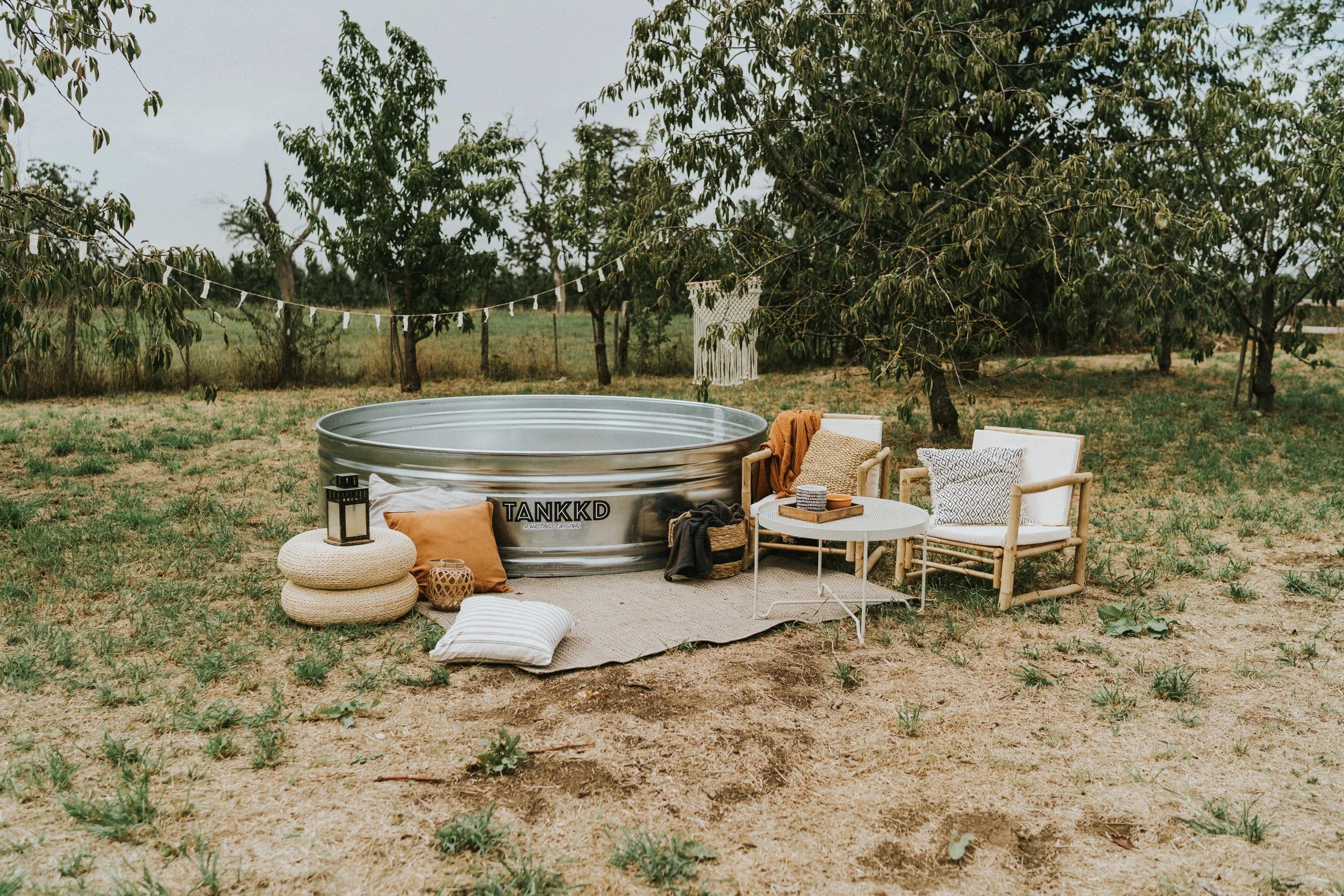 Outdoor scene with a metal tub, two white chairs with cushions, a small round table, and decorative pillows and lanterns on a beige rug in a grassy yard with trees in the background.