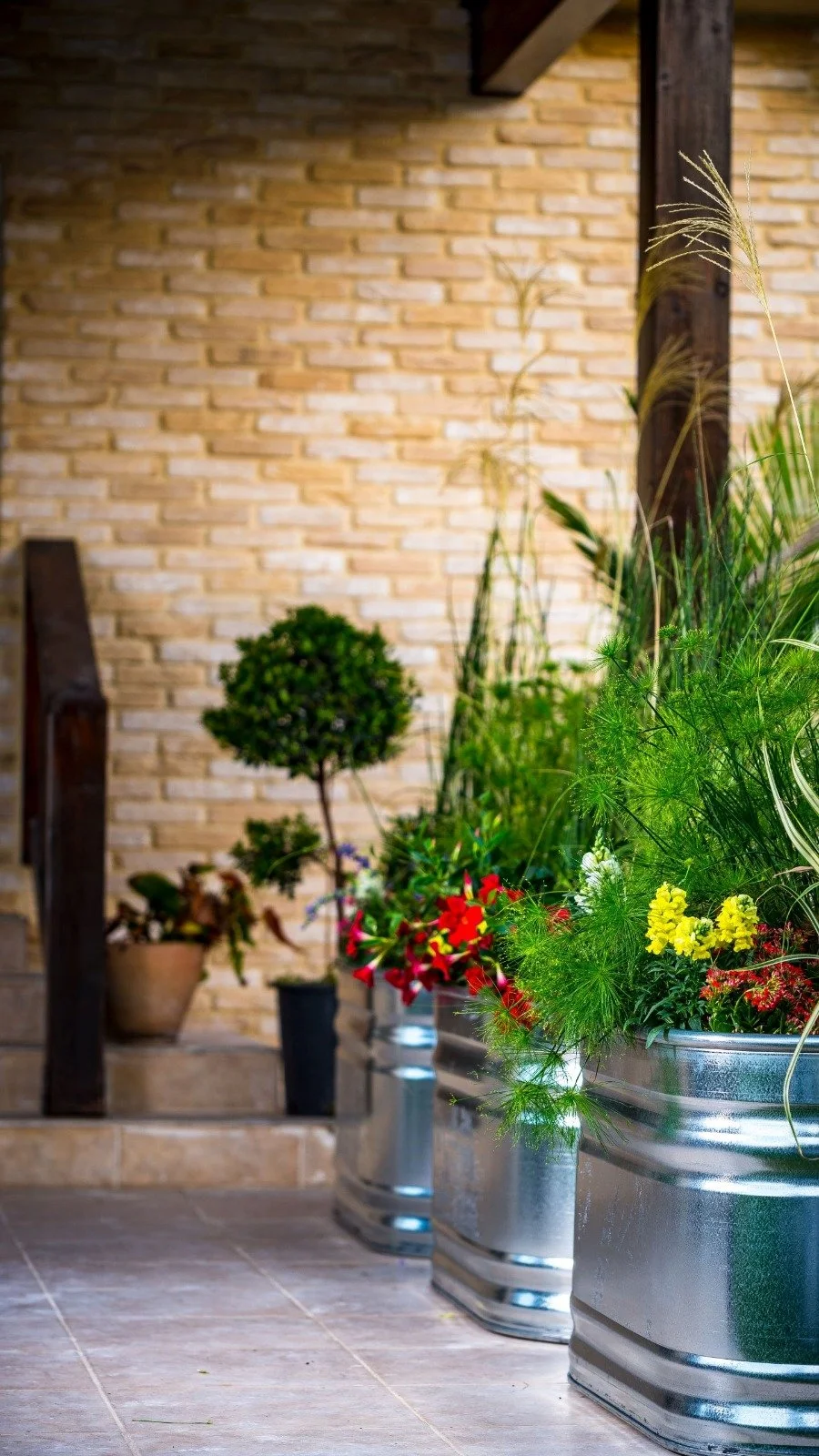 Decorative potted plants and flowers on tiled floor beside a brick wall and wooden railing in a Stock Tank