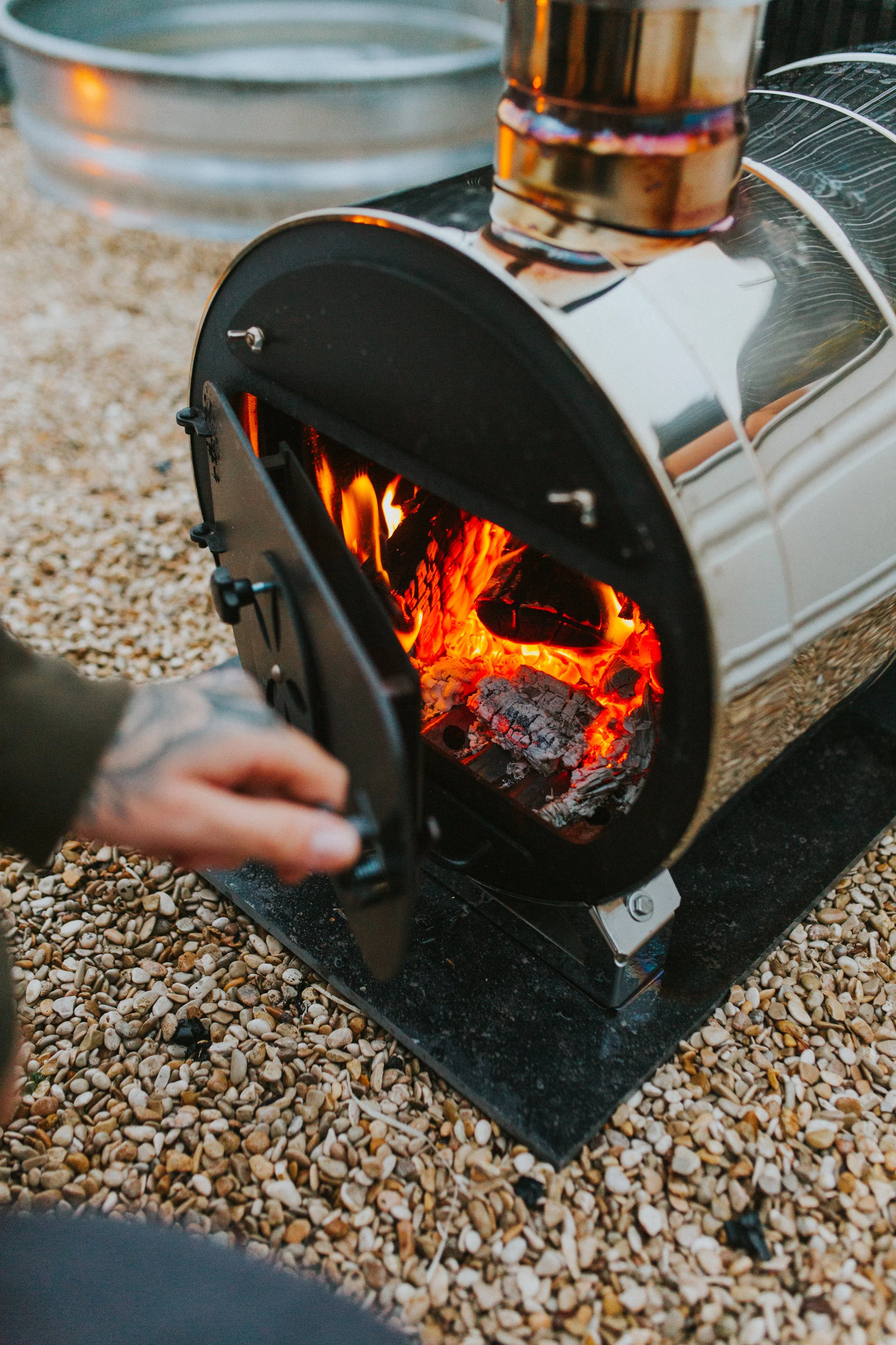 A person opening a small outdoor wood-burning stove with glowing embers inside, on a gravel surface.