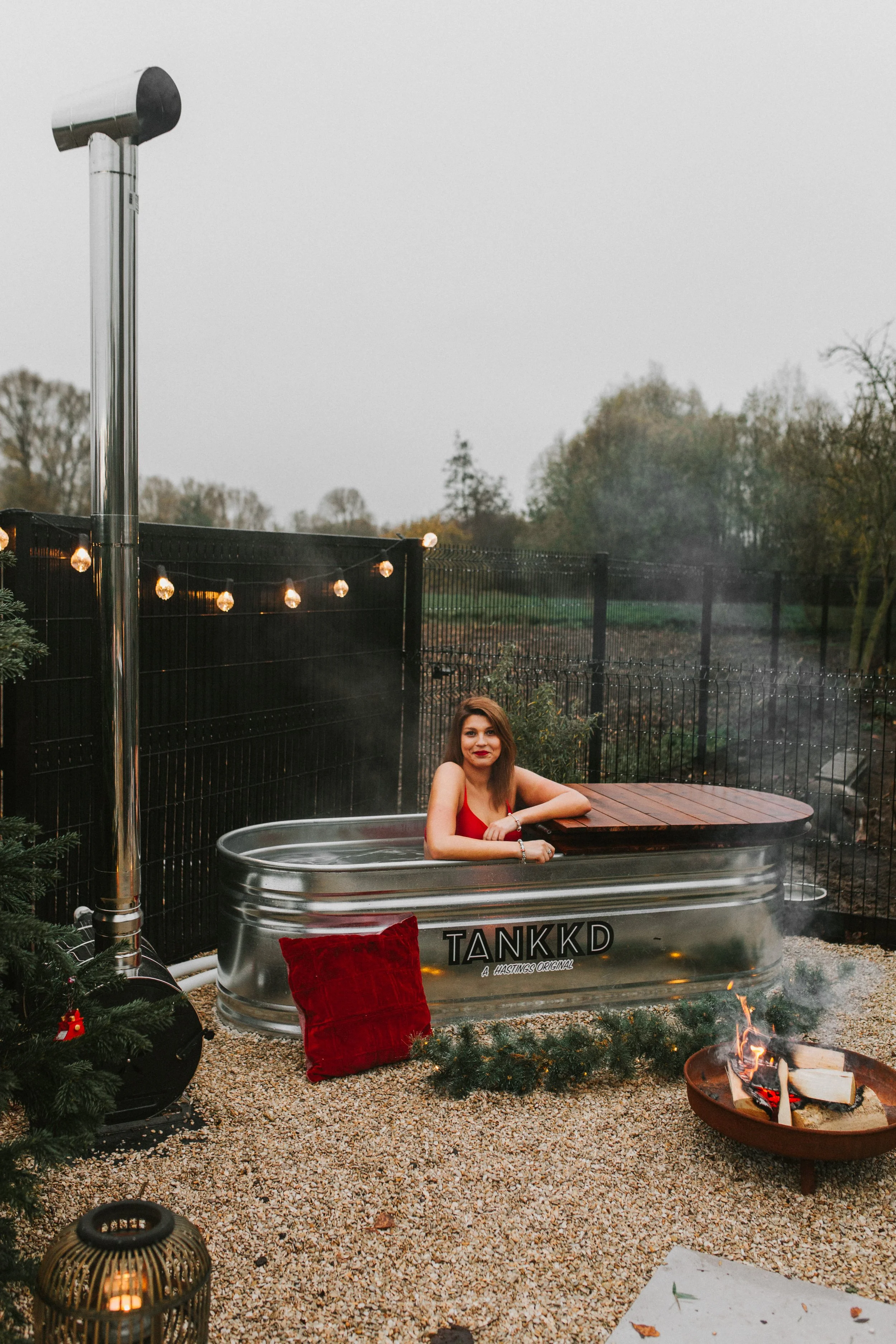A woman soaking in a metal hot tub outdoors during the evening, with string lights, a fire pit, and a Christmas tree nearby.