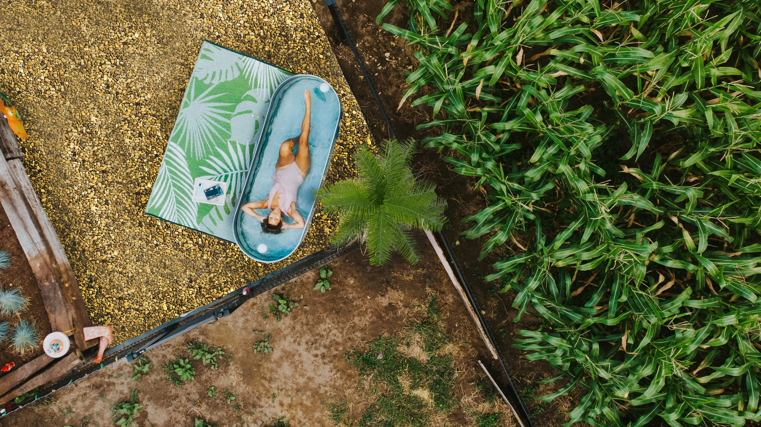 An aerial view of a woman in a pink swimsuit lying in a round stock tank pool , surrounded by green plants and fenced garden beds.