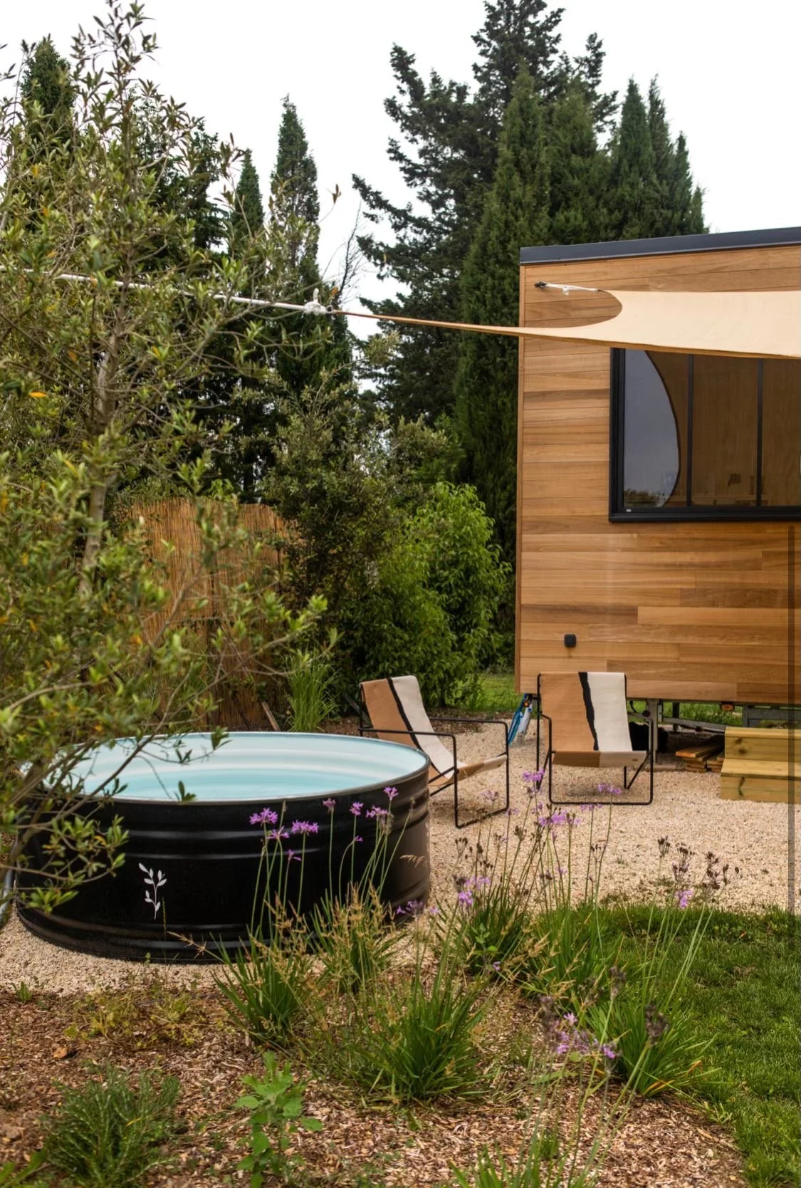 A backyard with a round black hot tub, two beige outdoor chairs, a wooden building with large windows, and lush green trees and flowering plants.