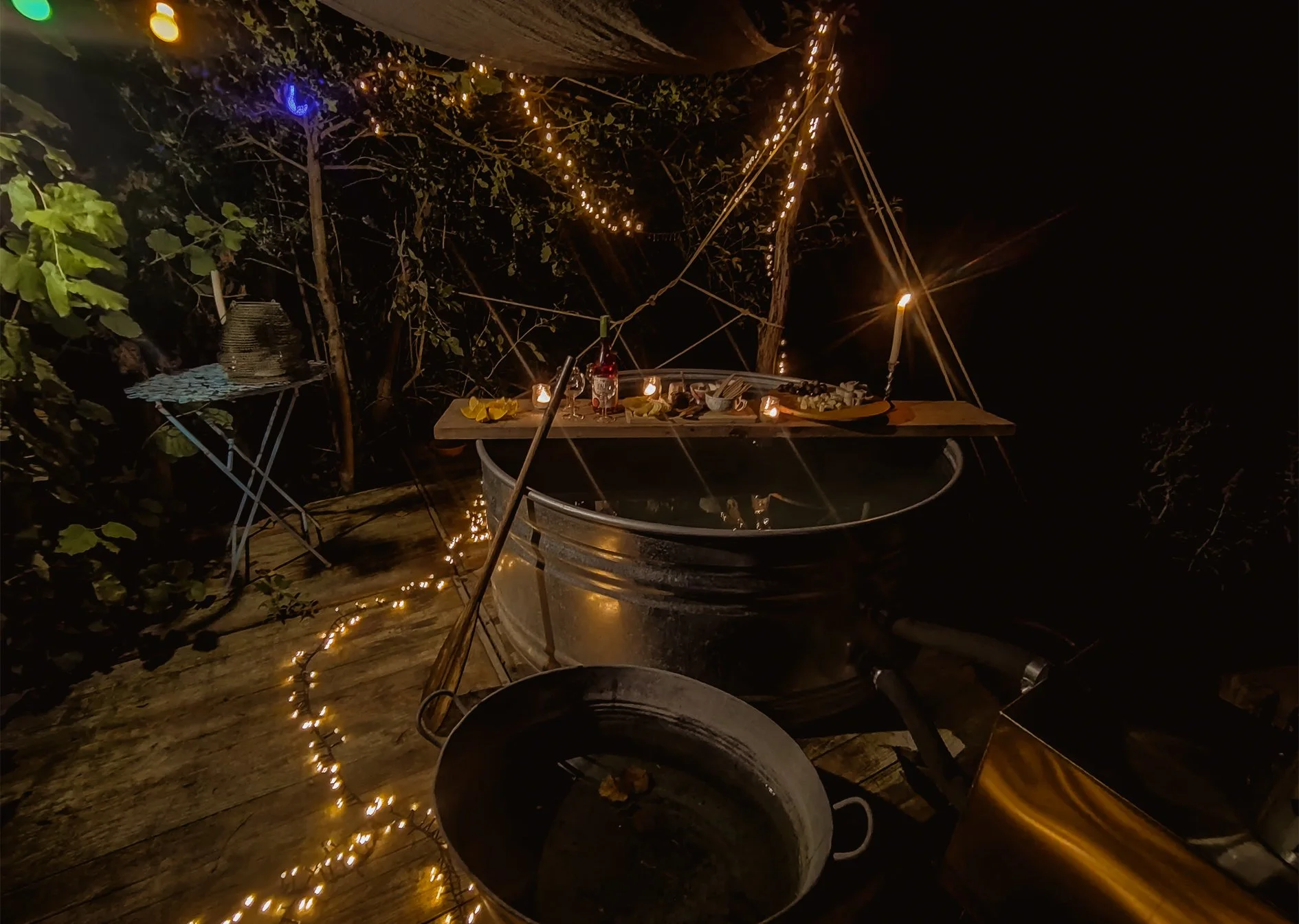 Outdoor nighttime scene with a rustic hot tub, a wooden table with candles, drinks, and snacks, surrounded by string lights and greenery.