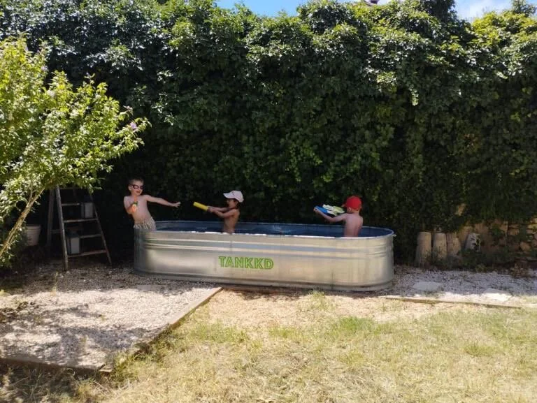 Three children playing in a Stock tank pool outdoors, with one wearing sunglasses, another wearing a cap, and a third wearing a red hat, surrounded by lush green bushes.