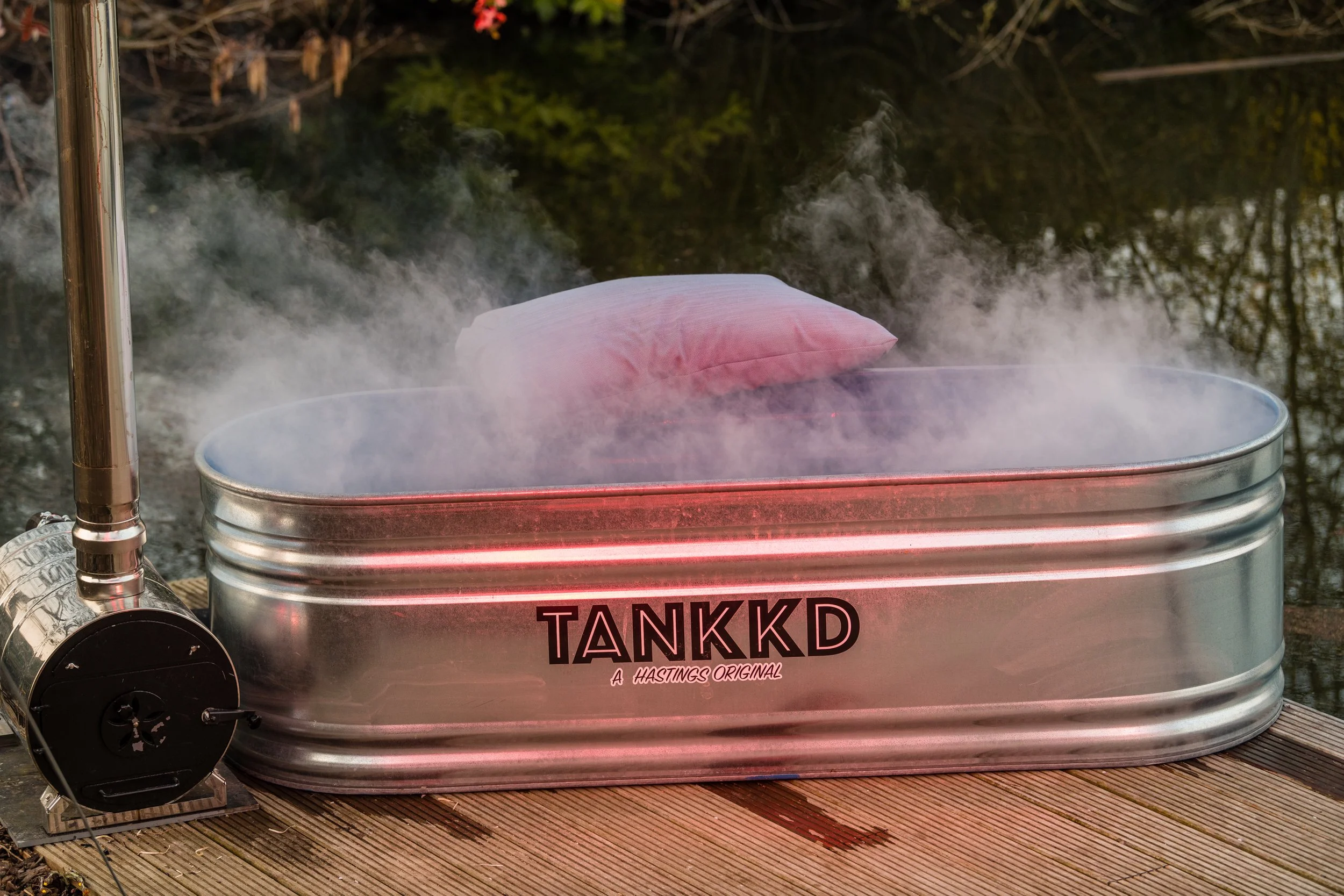 A metal tub labeled 'TANKKD' with steam rising from hot water and a pink pillow floating inside, outdoors on a wooden deck.
