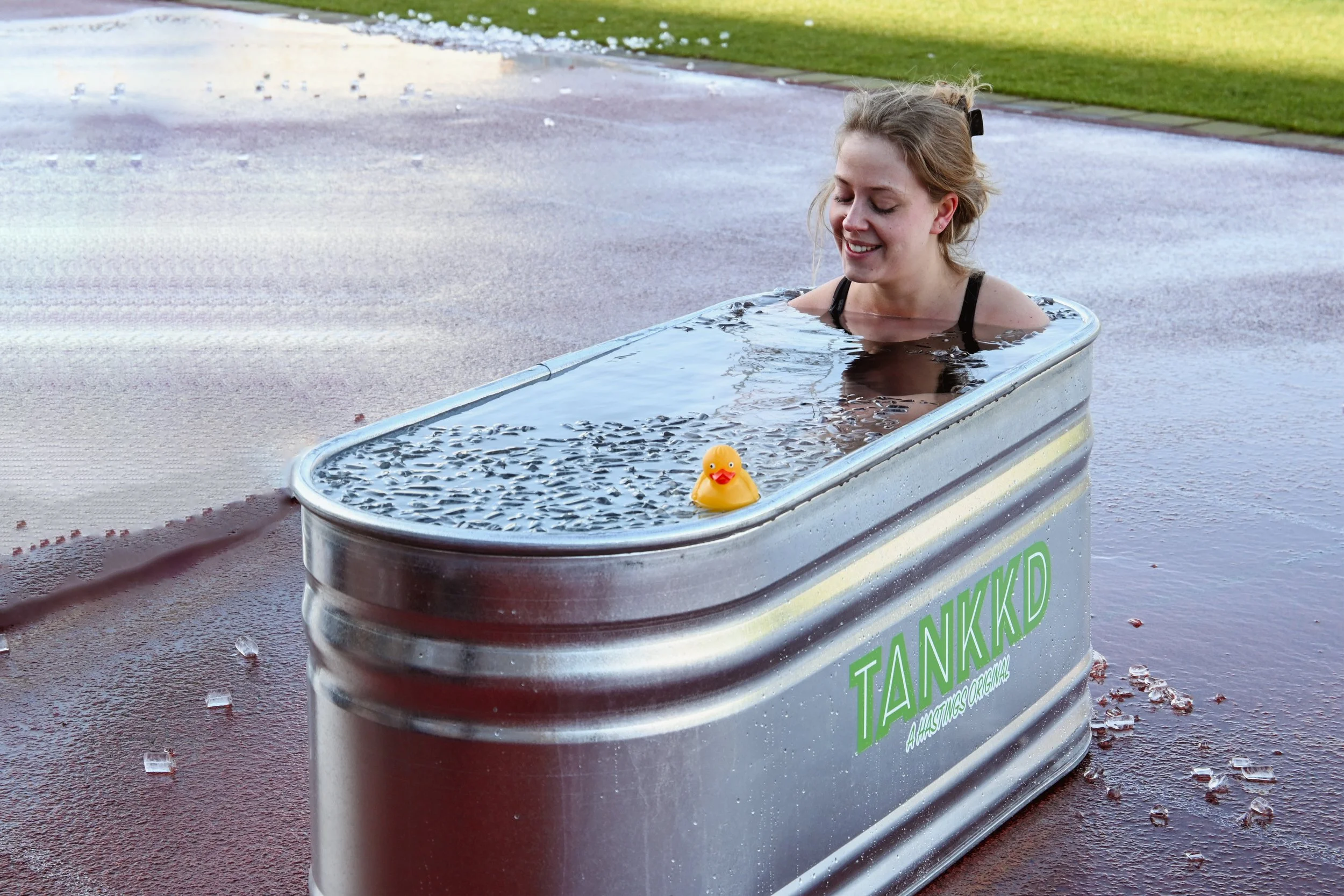 A woman soaking in a large metal tank labeled 'TANKKD' with a rubber duck floating on water inside.