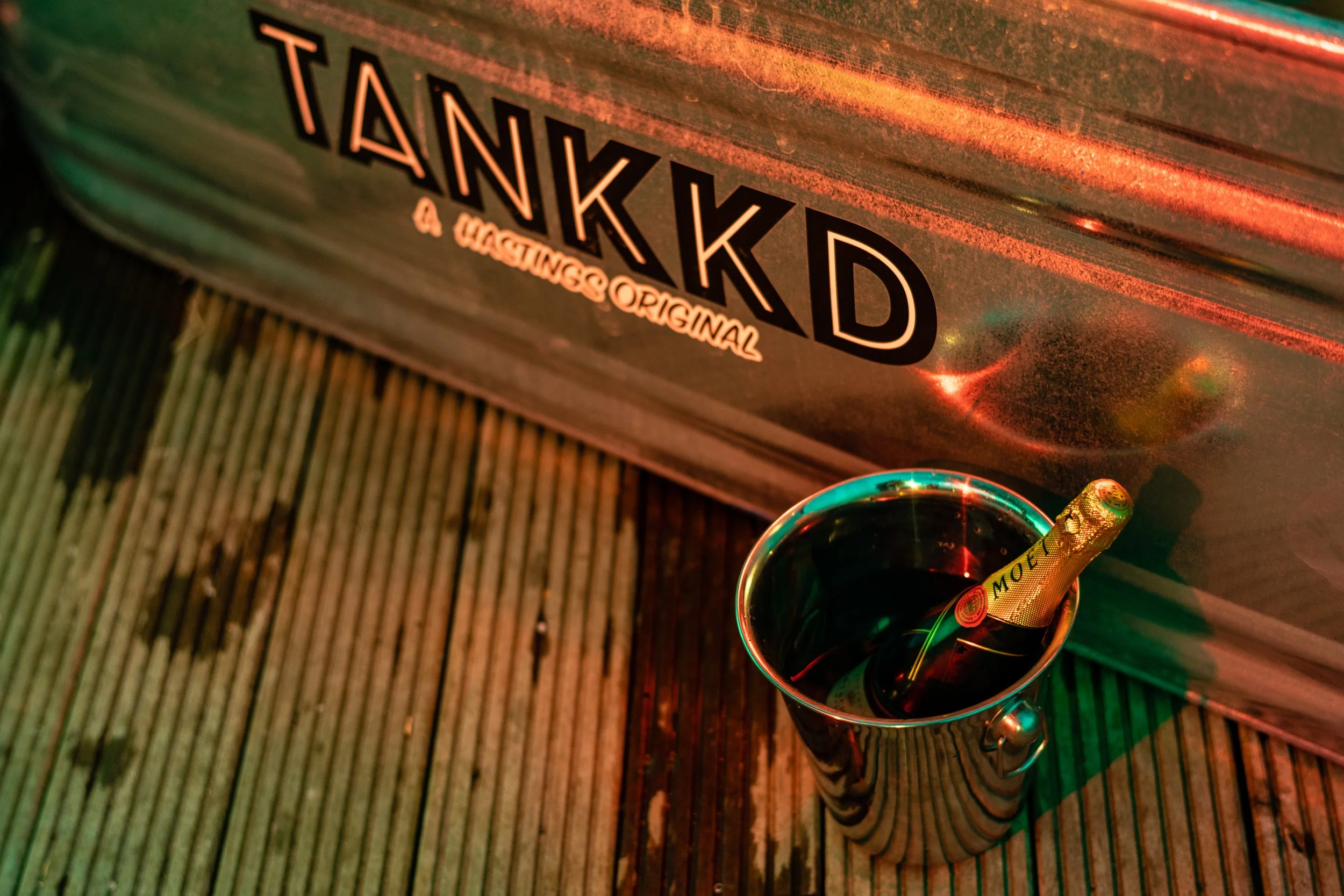 A mirror with a champagne bottle inside, placed on a wooden deck next to a metal storage box with the word "TANKKD" printed on it.