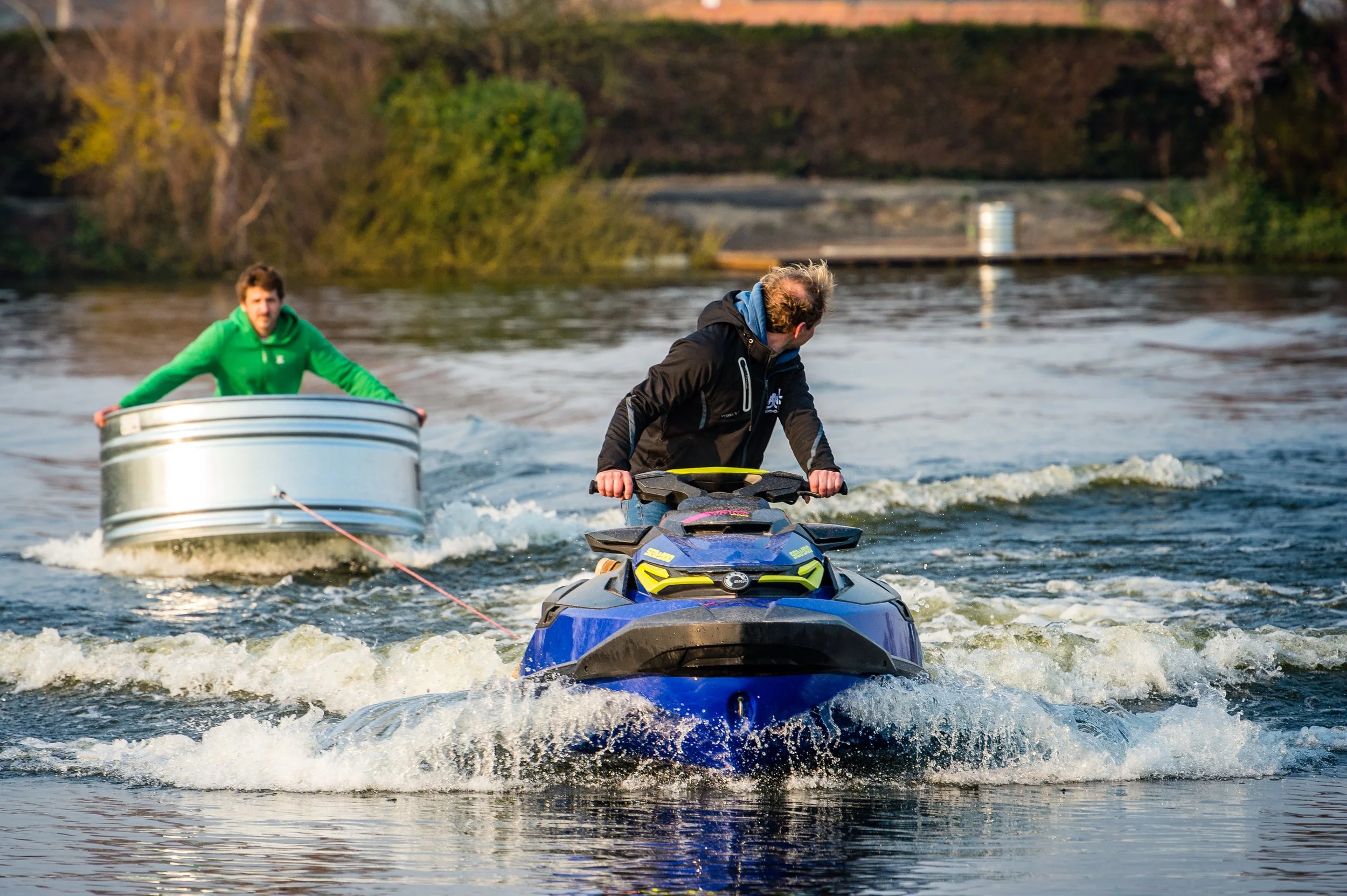 Two men riding a blue jet ski with a person sitting in a metal tub behind them on a river, with trees and a concrete structure in the background.