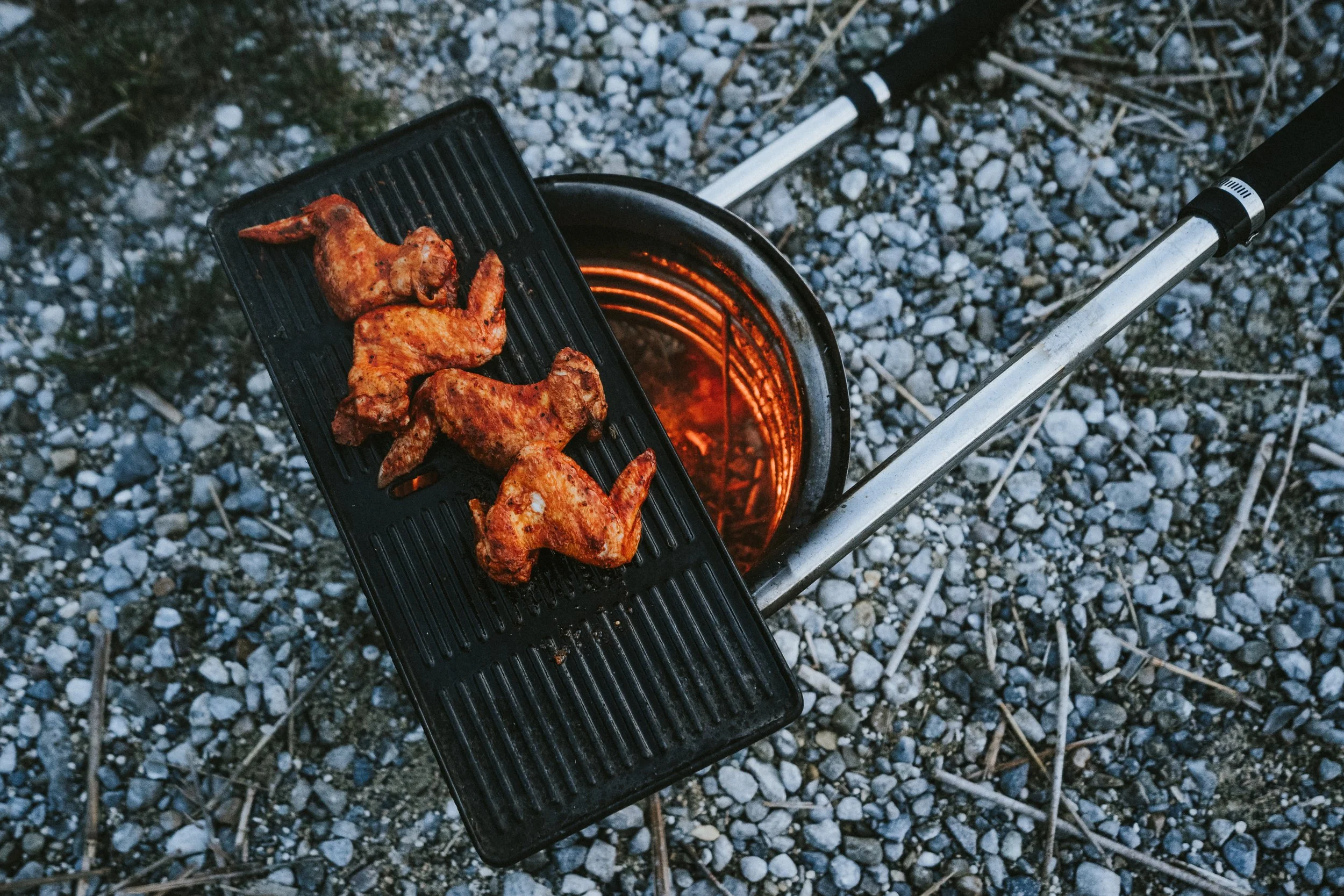 Four cooked chicken wings on a black grill rack placed over a small open grill on rocky ground.