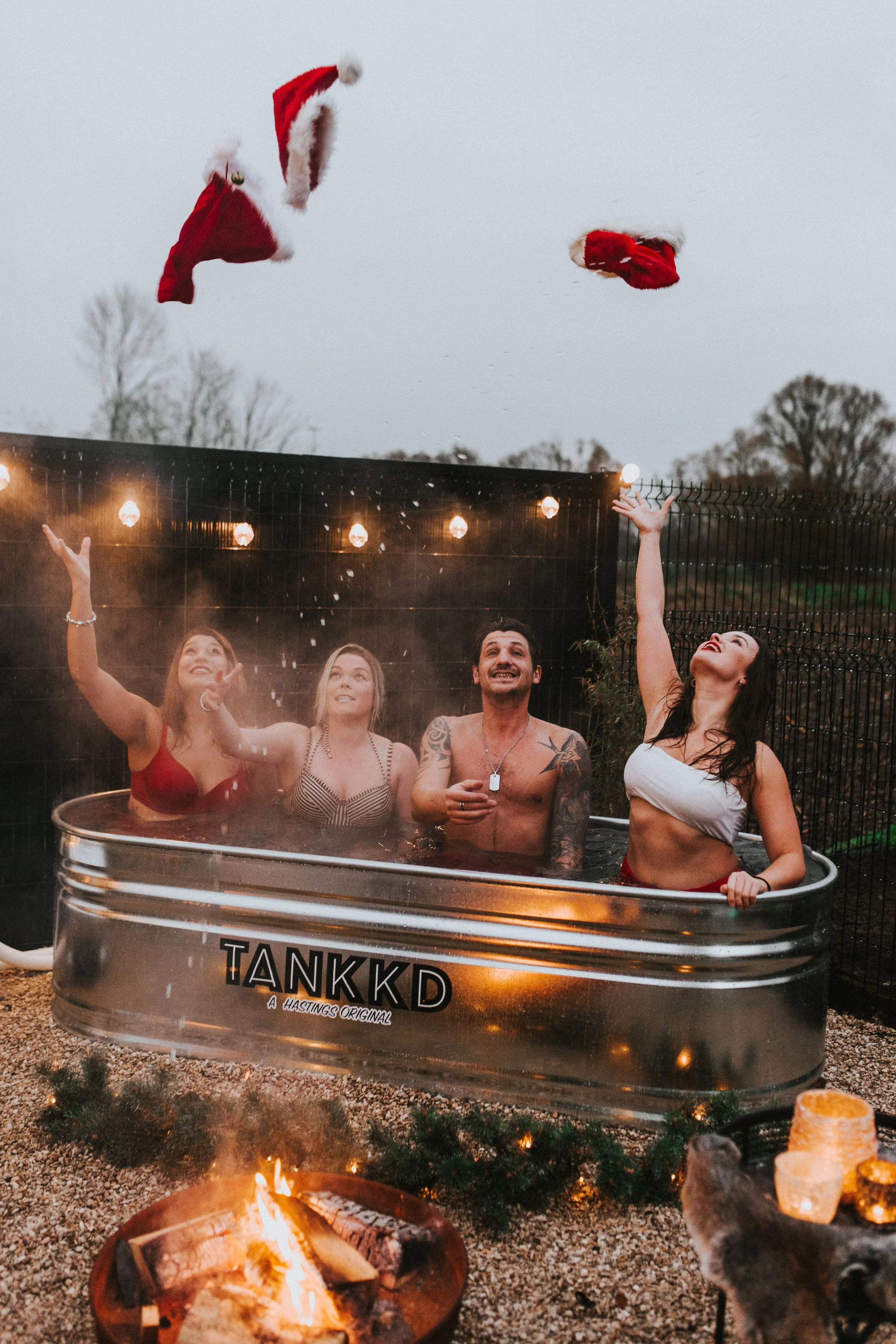 A group of four people in a Stock tank pool outdoors with holiday lights and a Christmas fire pit, tossing Santa hats into the air.