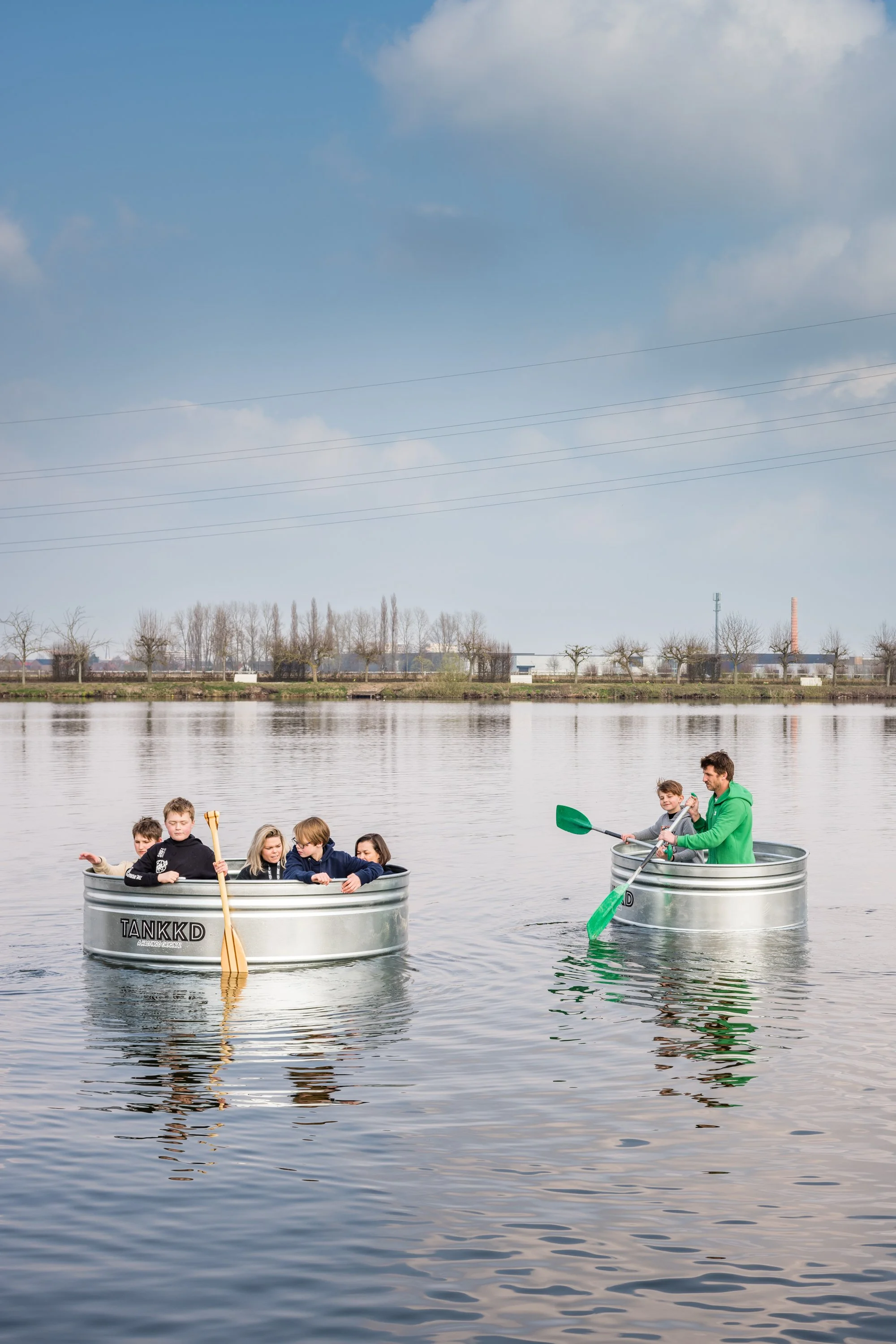 Children and an adult in two metal tubs floating on a body of water, using paddles to navigate, with a cityscape in the background.
